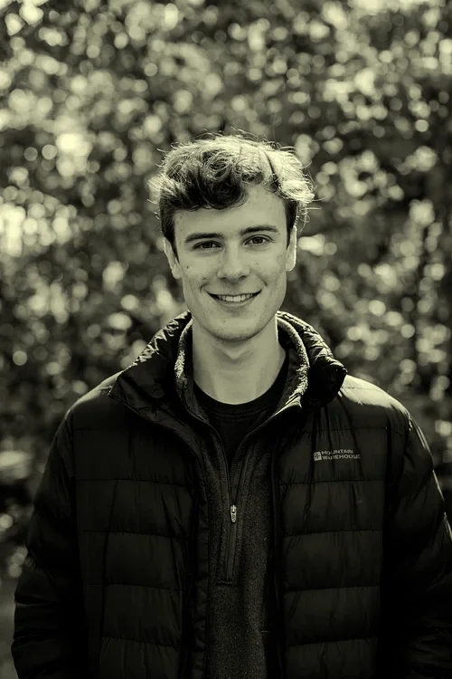 A young man smiling outdoors, wearing a jacket, with blurred trees in the background.