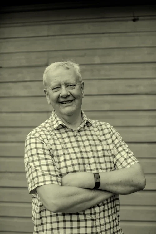 A smiling elderly man with short light-colored hair, wearing a checkered shirt and a watch, standing with his arms crossed in front of a wooden wall.