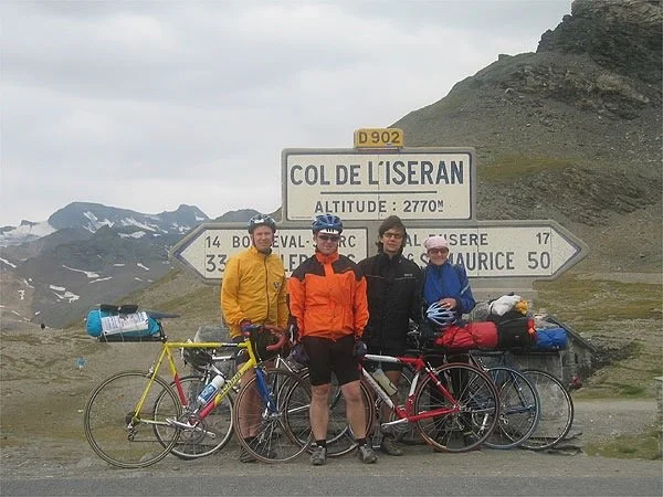 Vier Radfahrer in sportlicher Kleidung mit Fahrrad vor einem Wegweiser bei Col de l' Iseran, im Hintergrund Berge und Schneefelder, bewölkter Himmel.