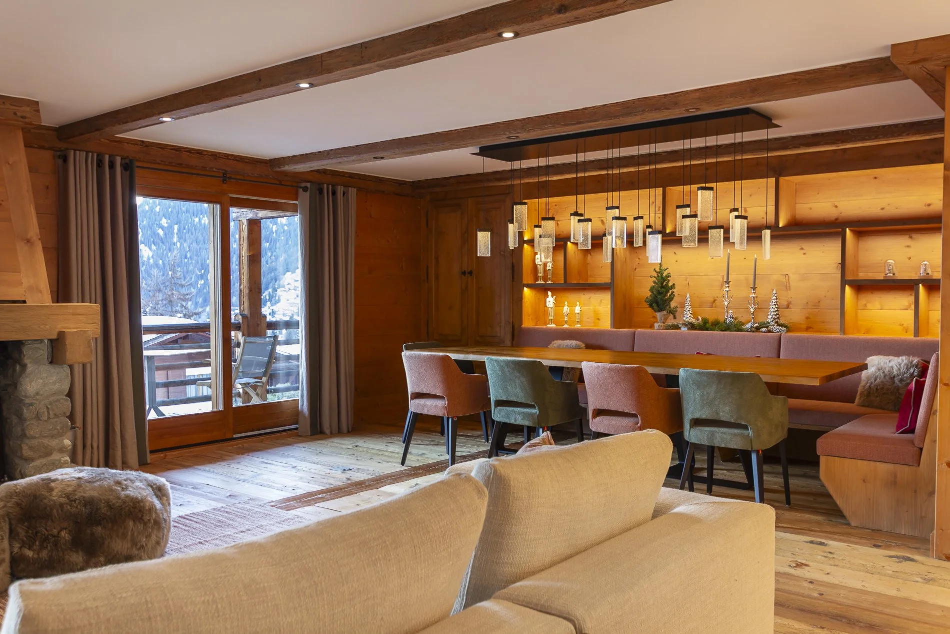 Cozy living room with wooden walls and ceiling beams, multi-colored chairs around a wooden table, decorated with holiday ornaments and small Christmas trees, large window with snowy landscape outside.