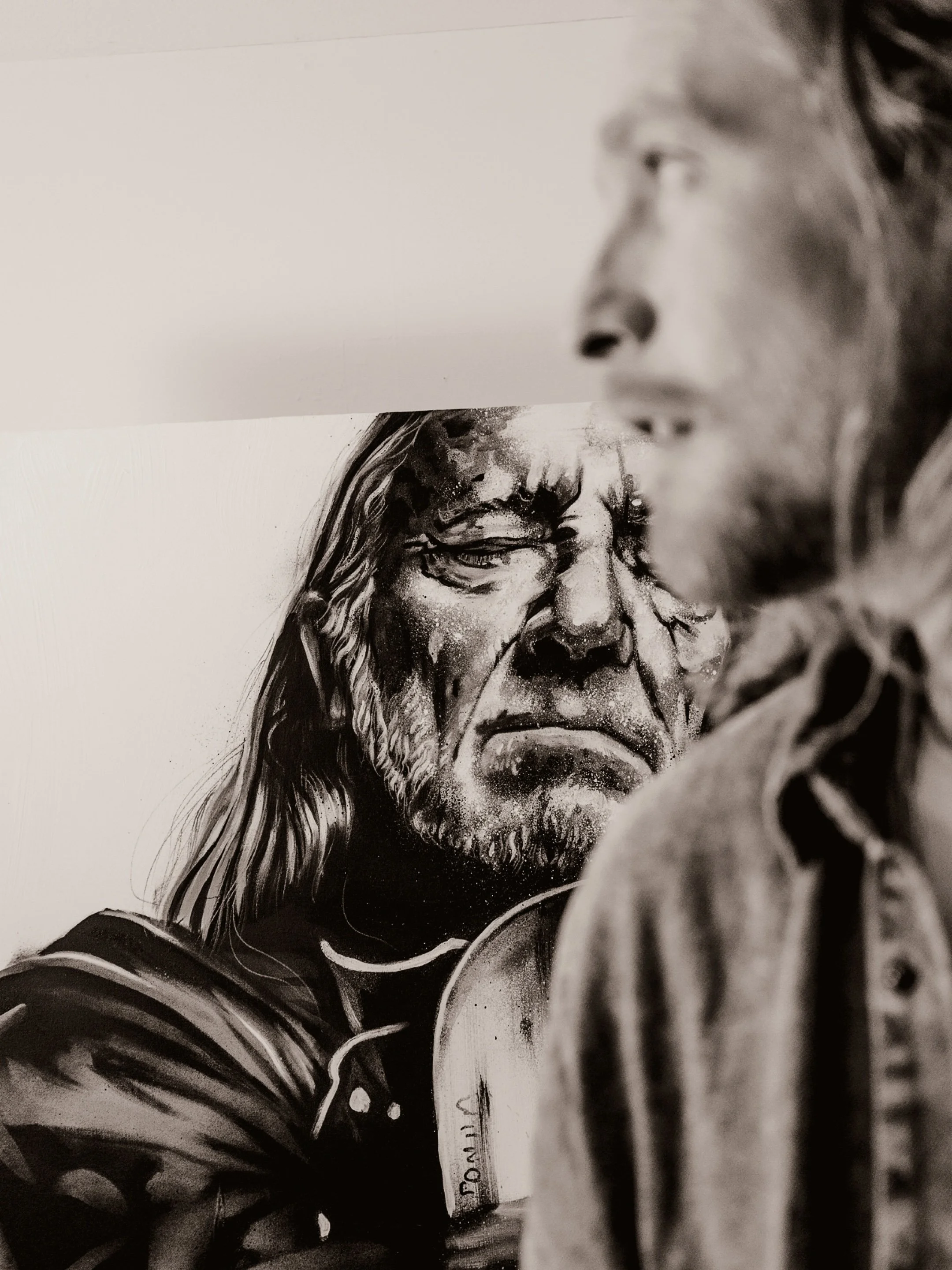 A woman with long hair and a beard, wearing a dark jacket, holding a cup, with a portrait of a person with long hair and a beard in the background.