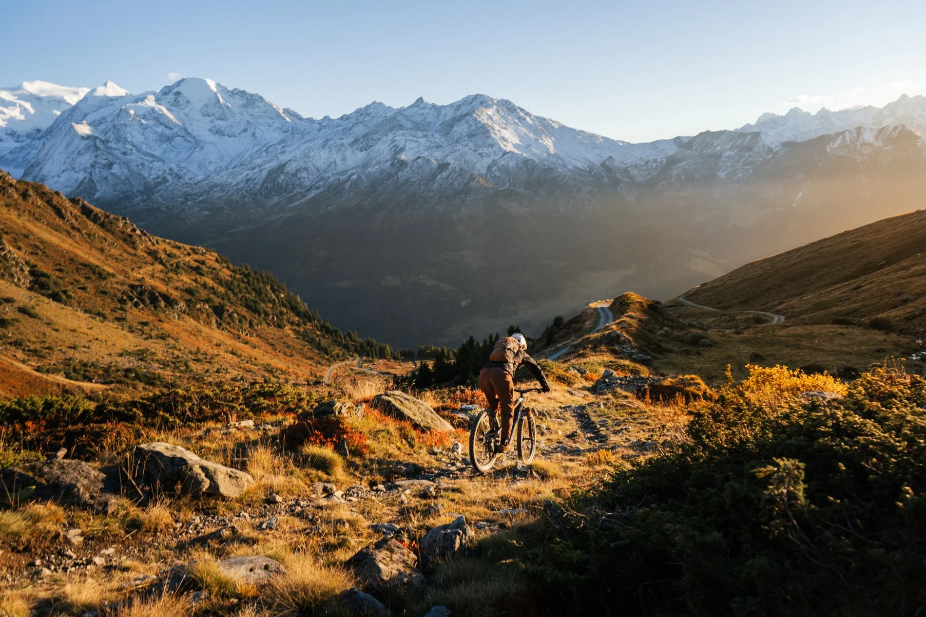 A person mountain biking on a trail in a mountainous landscape with snow-capped peaks and a clear sky in the background, during late afternoon or early evening.
