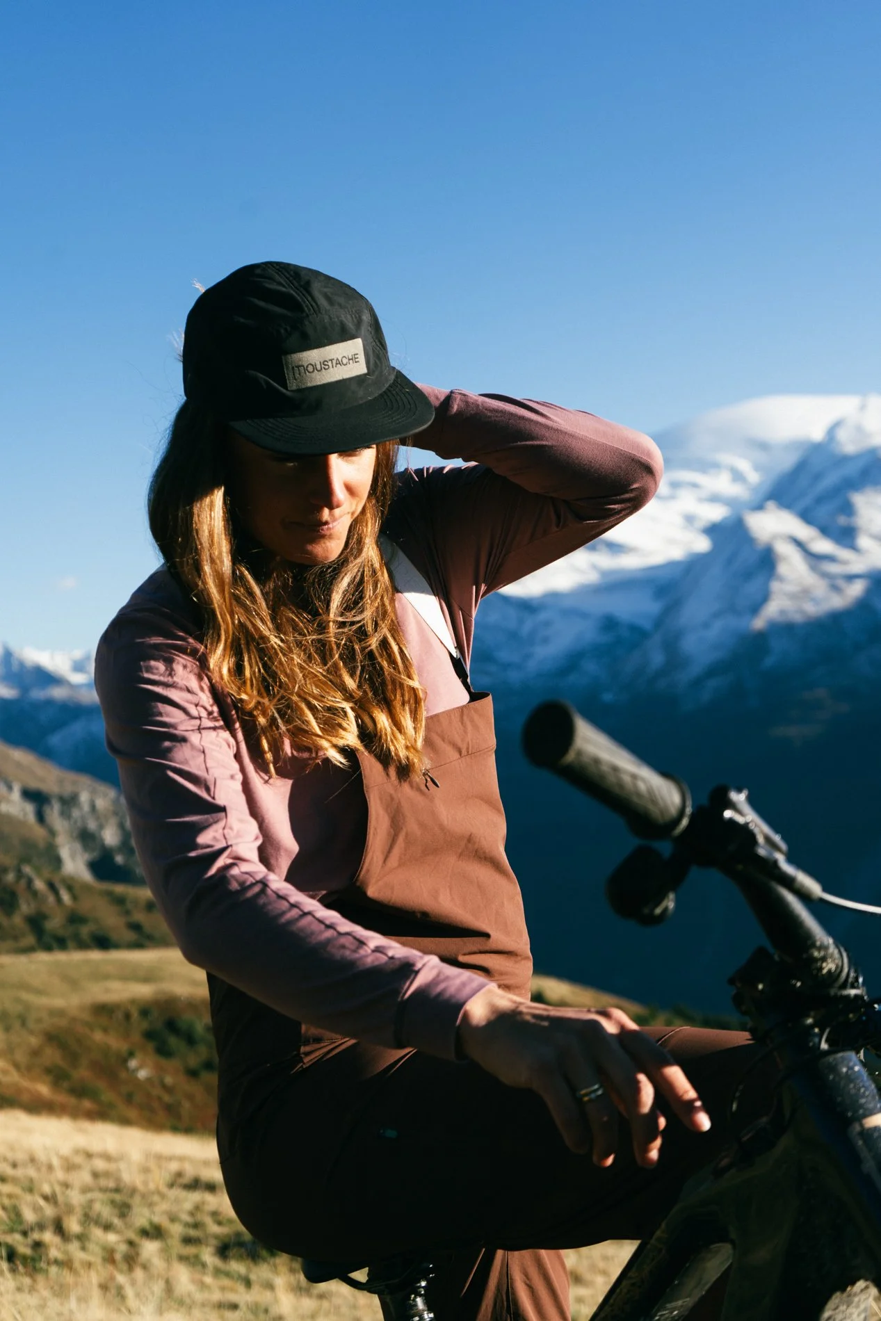 A woman wearing a black cap and brown jacket sits on a mountain bike with snow-capped mountains in the background under a clear blue sky.