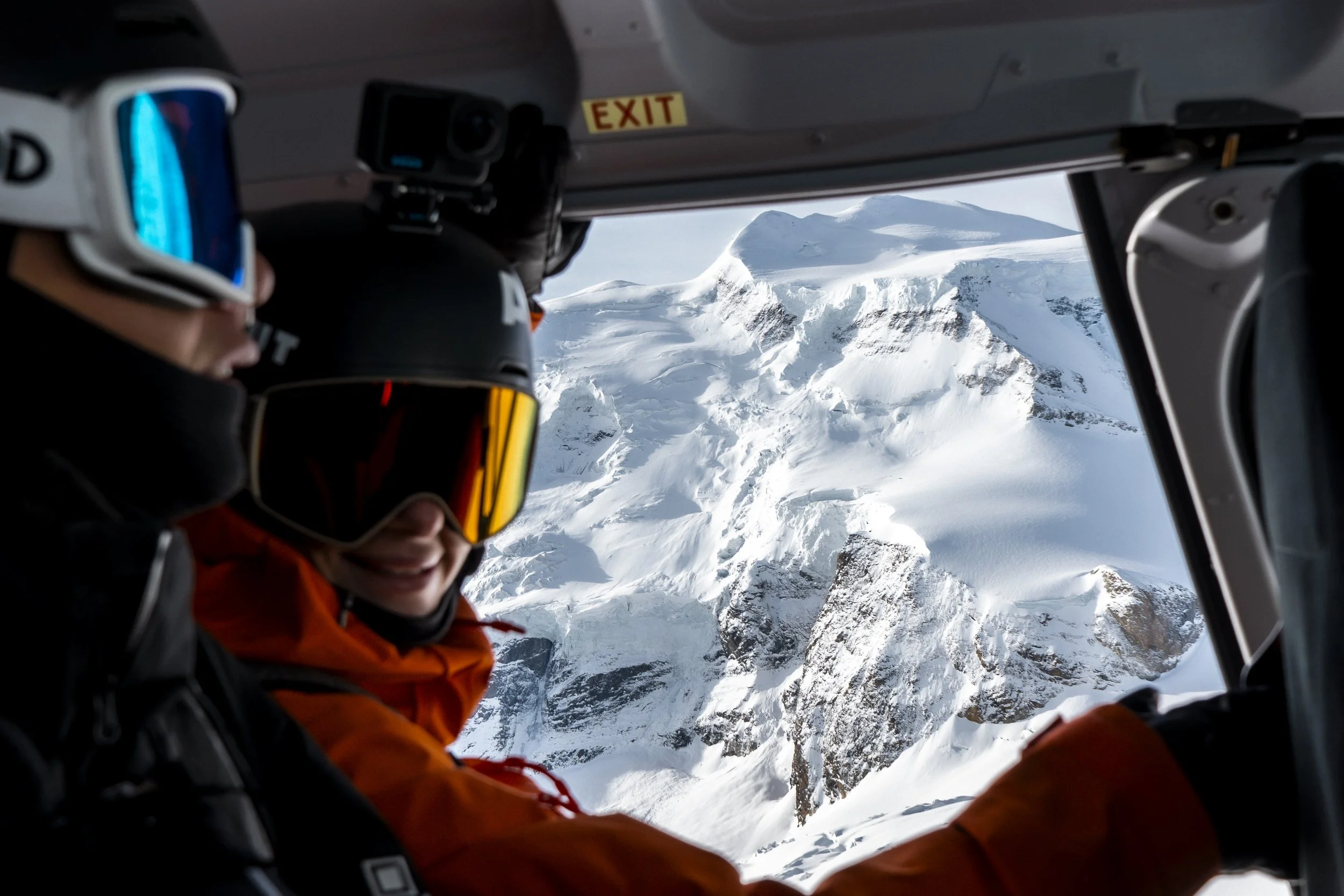 Two people inside a helicopter flying over snowy mountains, wearing helmets and winter gear.