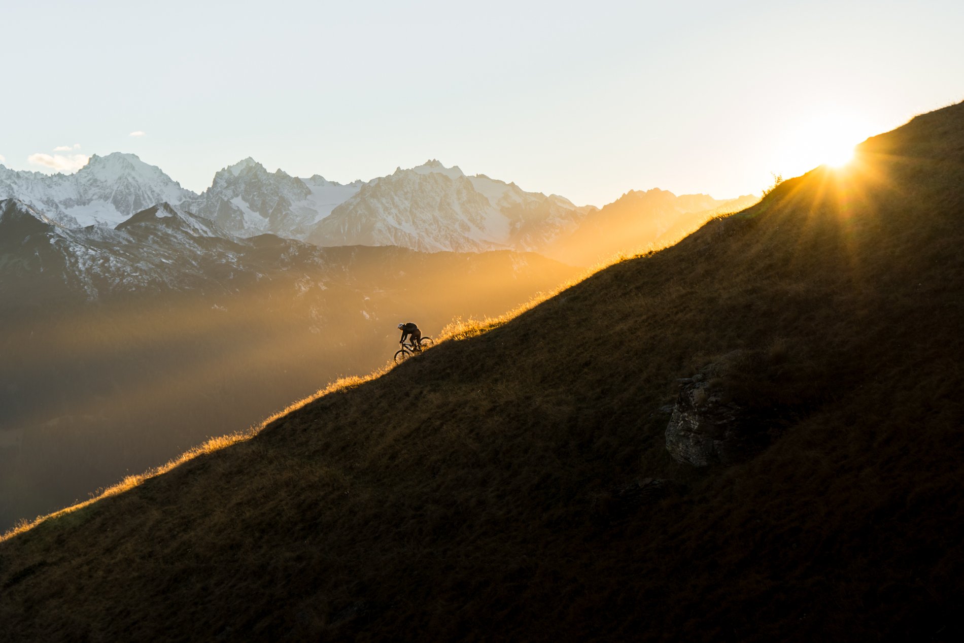 A cyclist riding uphill on a grassy mountain slope at sunset with snow-capped mountains in the background.