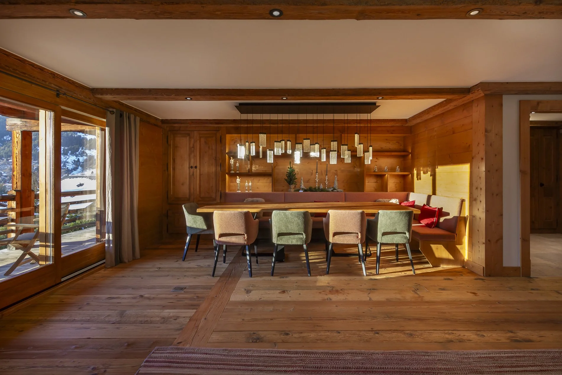 A cozy dining area with a large wooden table surrounded by multicolored upholstered chairs, illuminated by hanging pendant lights, with wooden panel walls and a sliding glass door showing snowy mountain scenery outside.