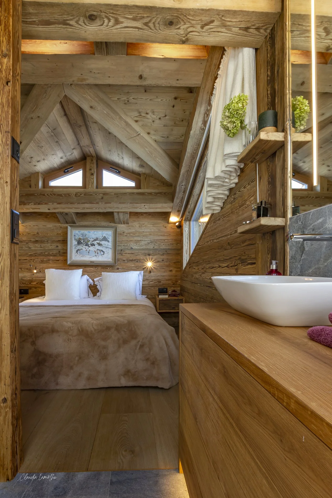 Cozy rustic bedroom with wood-paneled walls and ceiling, featuring a bed with white pillows, a painting above the bed, and a small sink with a red soap dispenser on a wooden cabinet, with a window partially covered by a curtain and decorative plants.