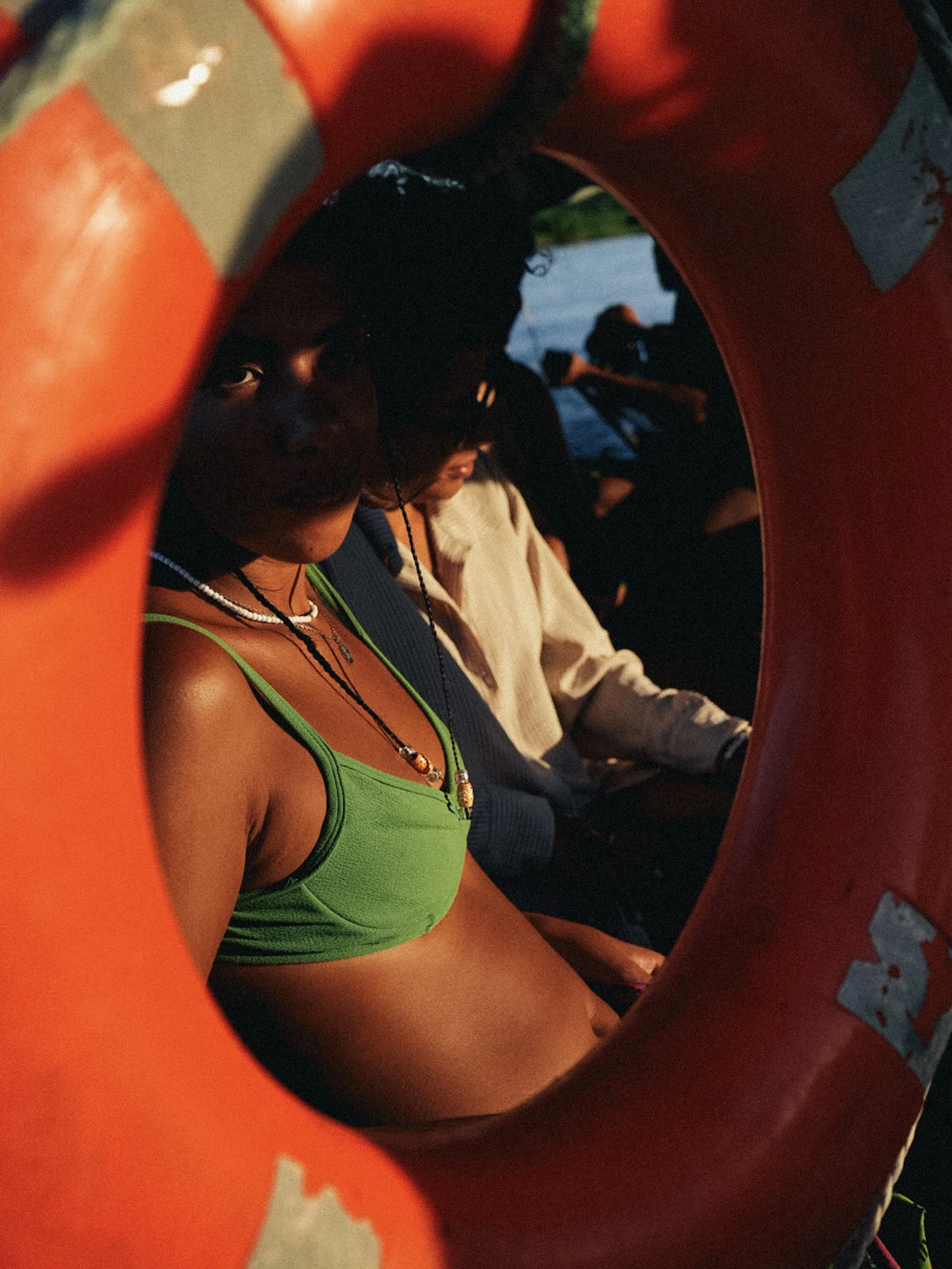 A person wearing a green top and multiple necklaces, partially visible through an orange life jacket or flotation device, looking towards the camera.
