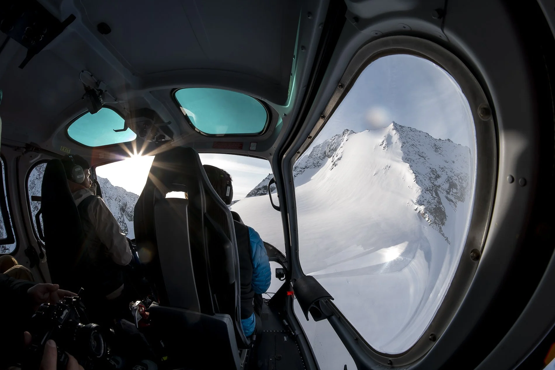 Inside a helicopter cockpit, pilots are flying over snowy mountain peaks with the sun shining through the windows.