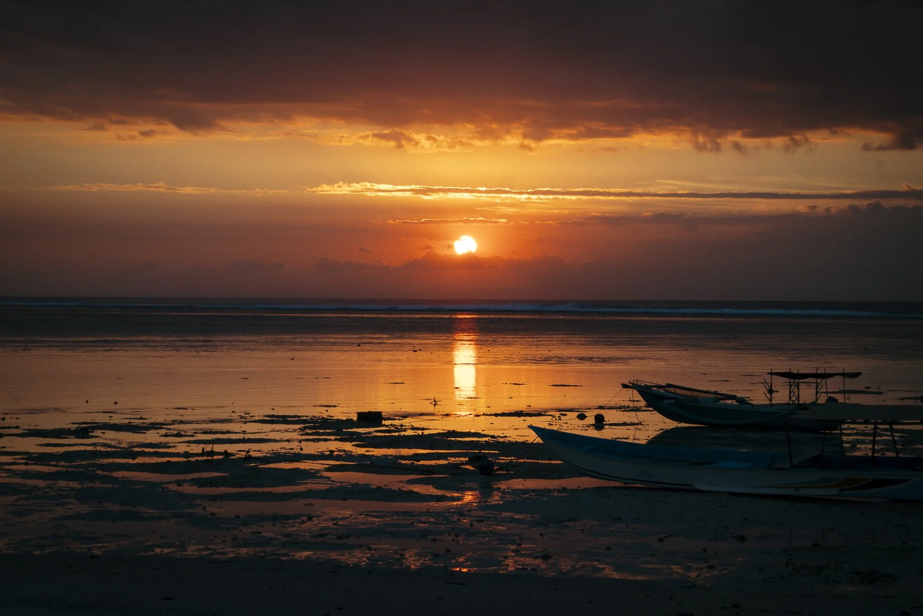 Sunset over the ocean with dark clouds in the sky, calm water reflecting the sky's colors, and three boats on the sandy shore.