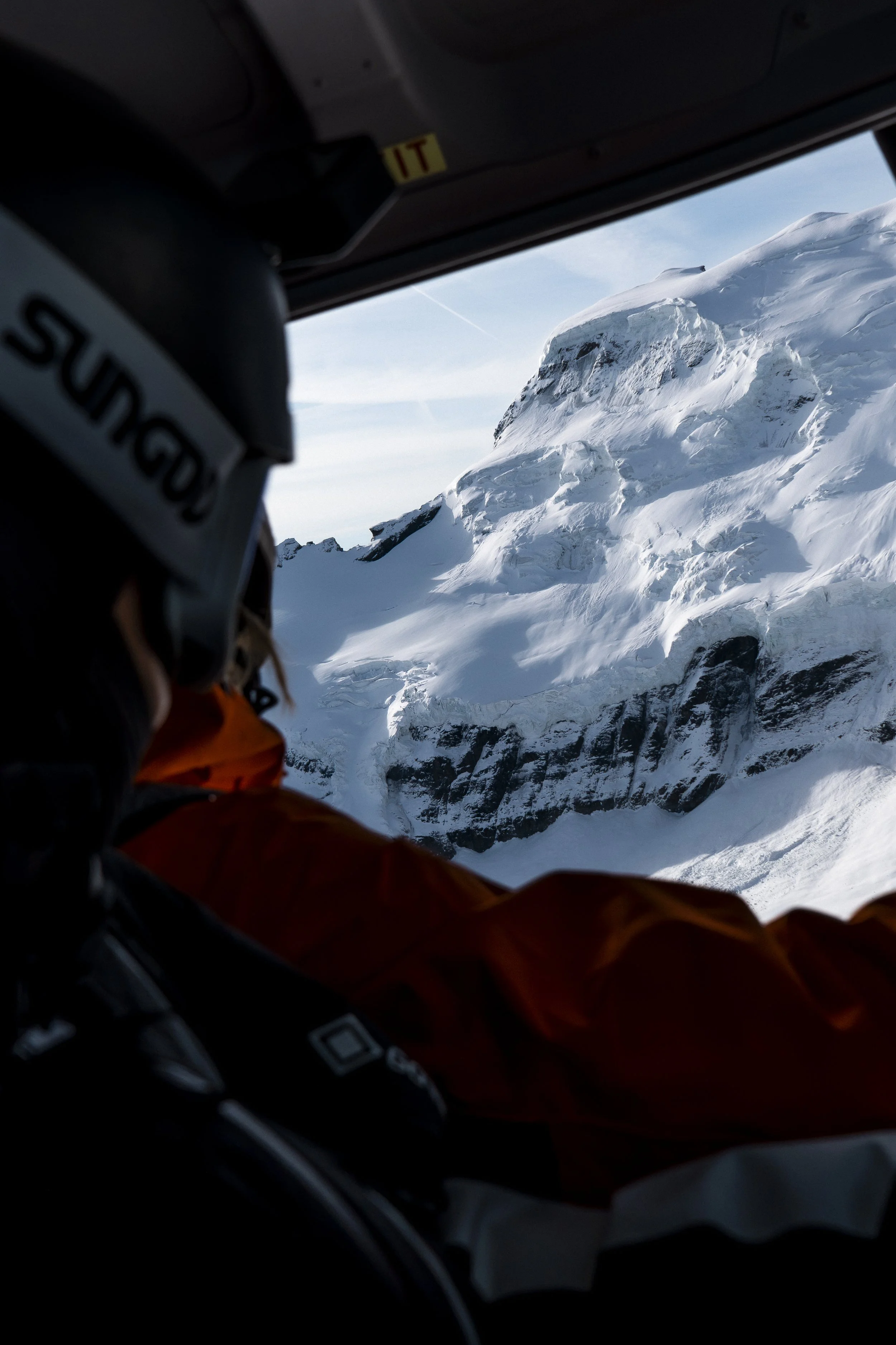 View from inside a helicopter looking out at a snow-covered mountain with jagged rocks. The helicopter's interior dark and partially visible.