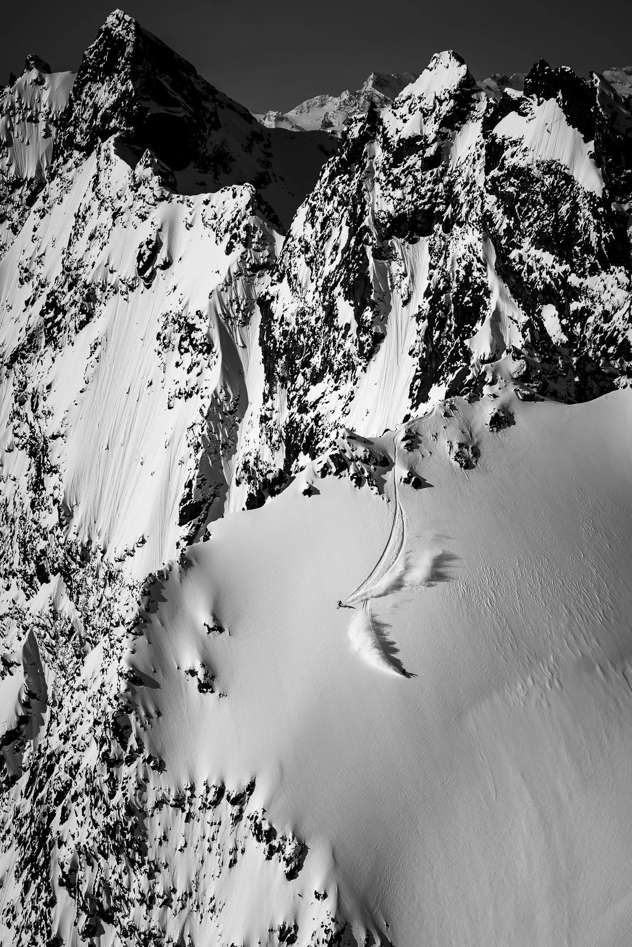 A black and white aerial view of snow-covered mountain peaks and a snowfield with ski tracks.