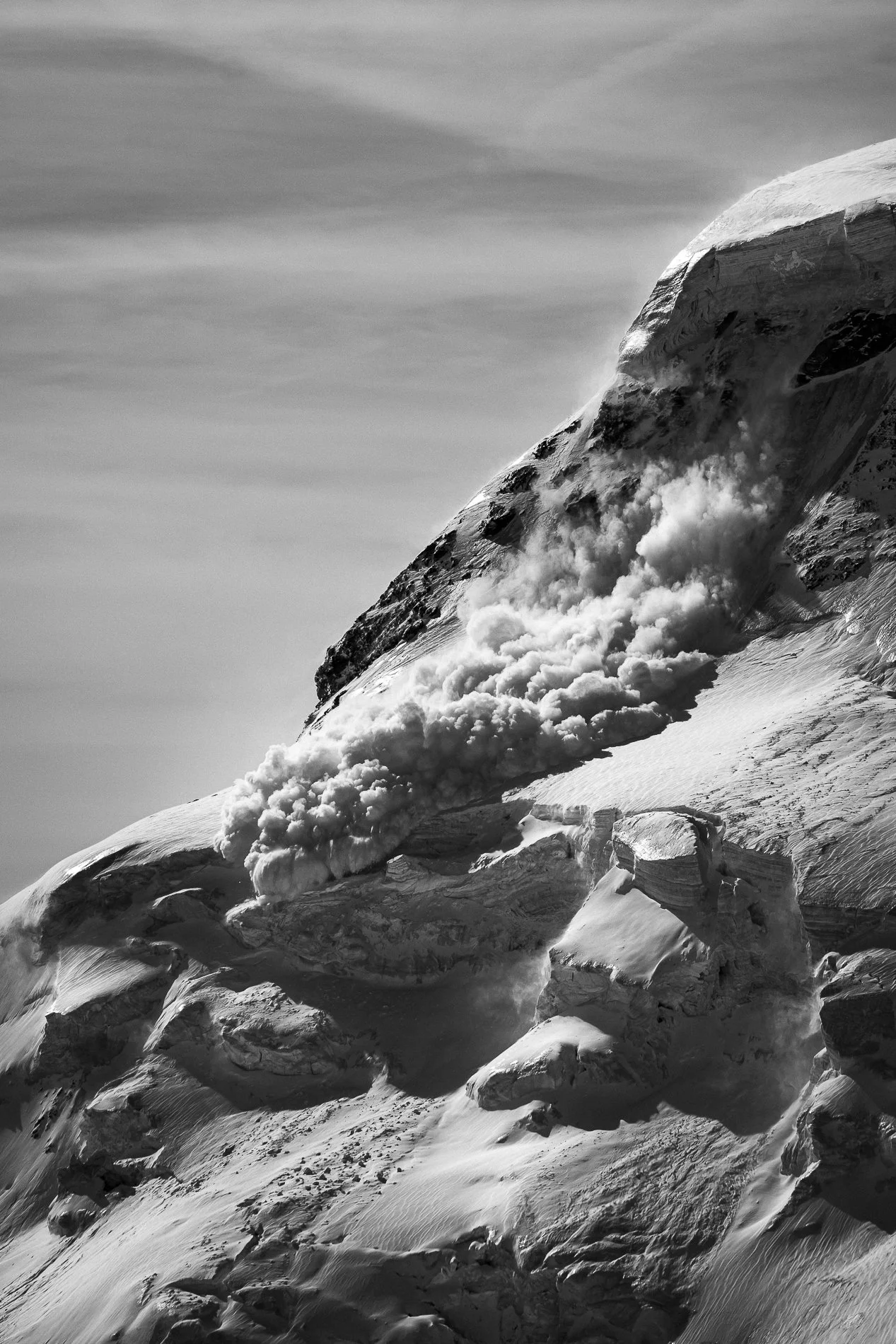 Black and white photograph of a snowy mountain with a snow-covered peak collapsing, creating a cloud of snow on the slope below.