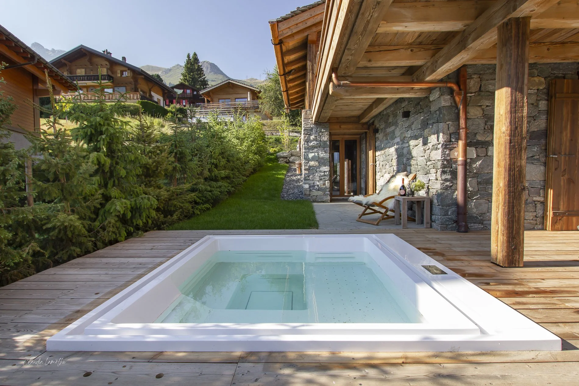 A backyard with a hot tub on a wooden deck, surrounded by green trees and shrubs. There are multiple wooden houses in the background on a hillside, with a mountain peak visible in the distance. The house in the foreground has a stone wall and a patio