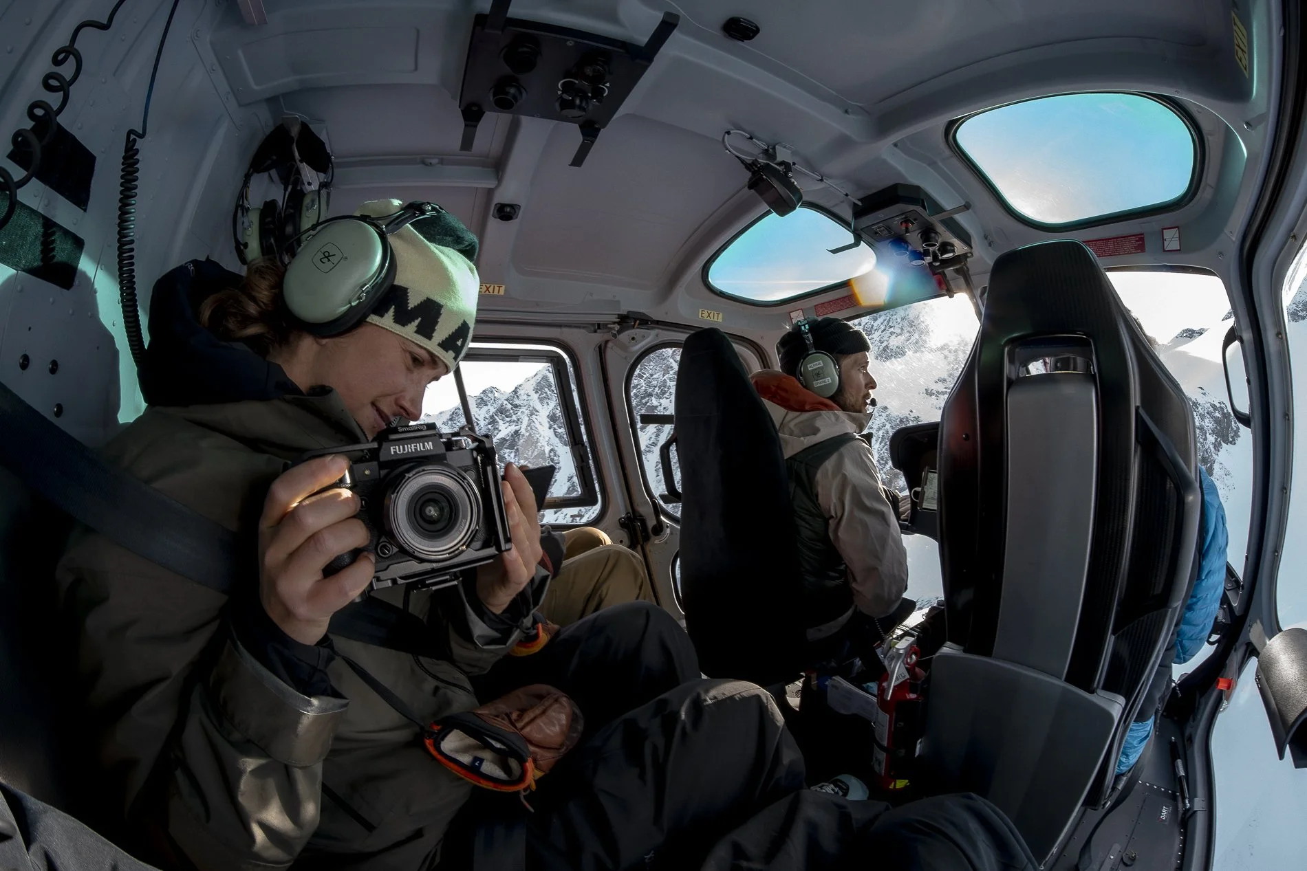 Inside a helicopter cockpit with two pilots, one woman holding a camera and a man sitting beside her, with snow-covered mountains visible through the windows.