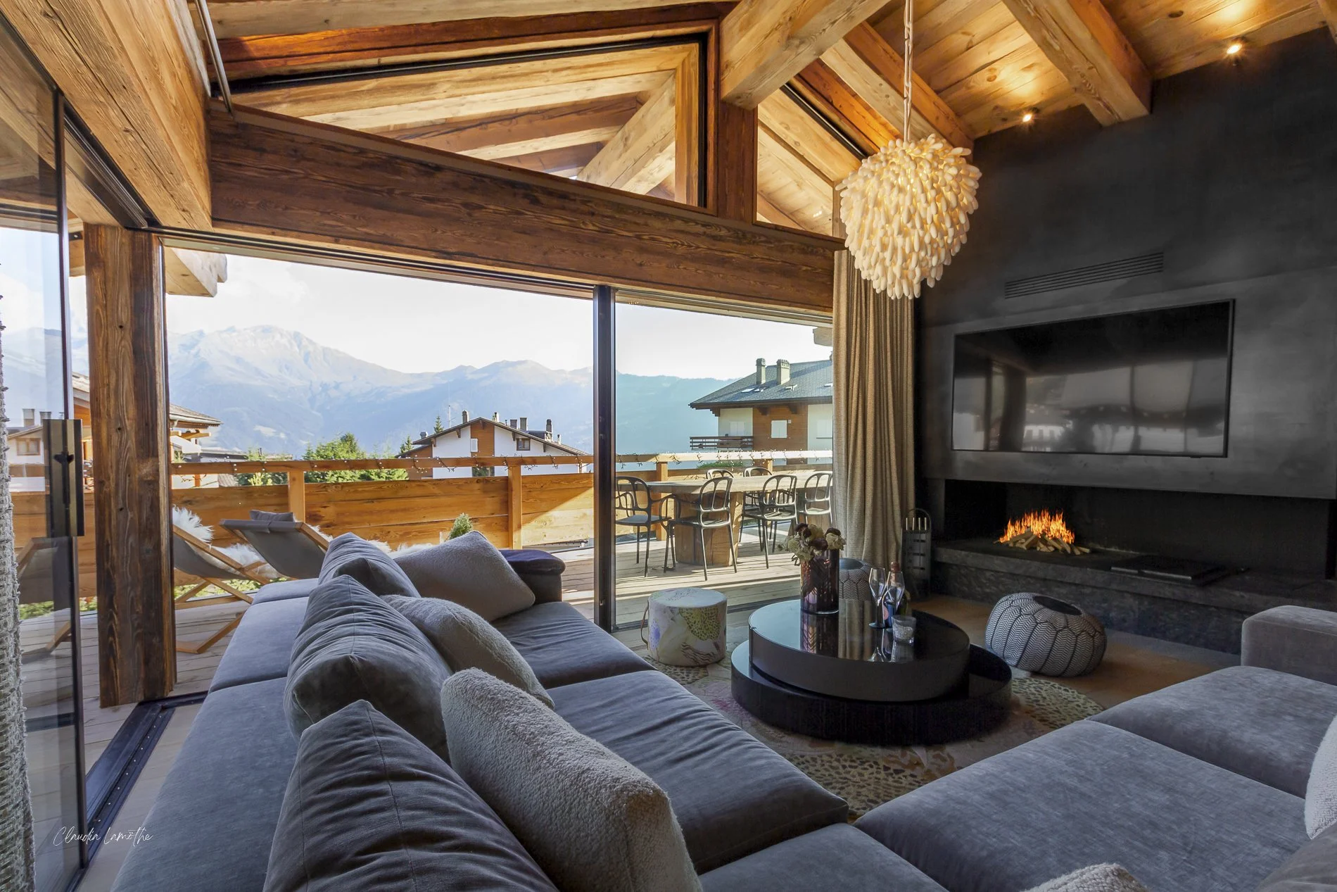 Living room with a gray sectional sofa, a black round coffee table, and a modern fireplace with a TV mounted above it. Large glass doors open to a balcony with outdoor seating, overlooking a mountain landscape.