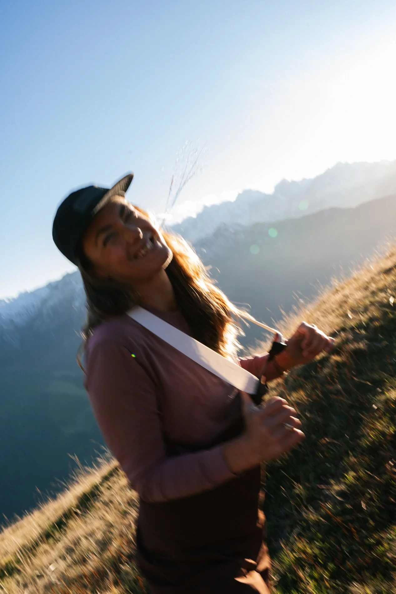 Woman smiling and jogging outdoors on a sunny day, wearing a cap and a pink long-sleeve shirt, with mountains in the background.