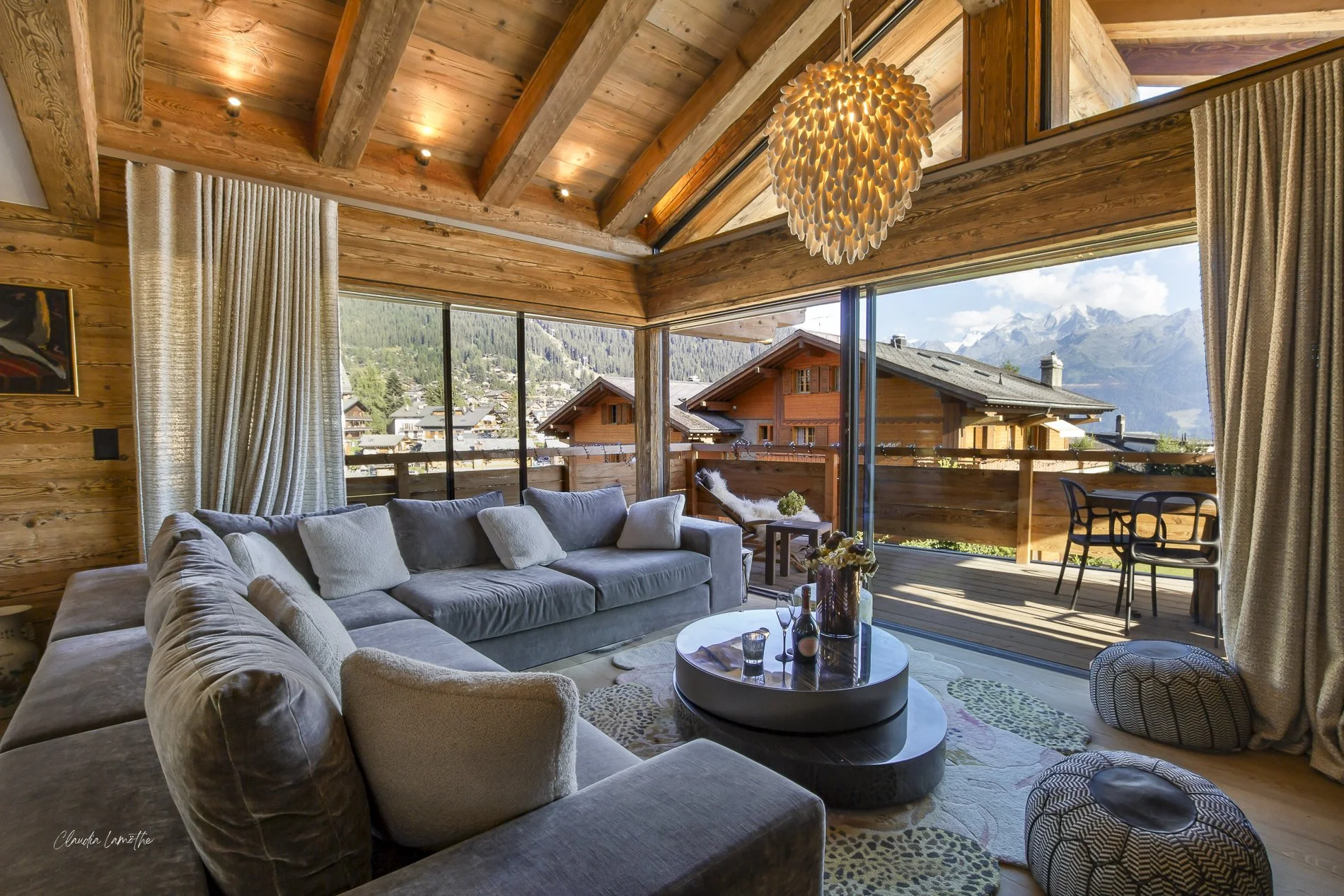 Living room with large beige sectional sofa, round black coffee table, and a view of a mountain landscape through floor-to-ceiling glass doors. Wooden ceiling with exposed beams and a coral chandelier, beige curtains, and outdoor balcony with chairs 