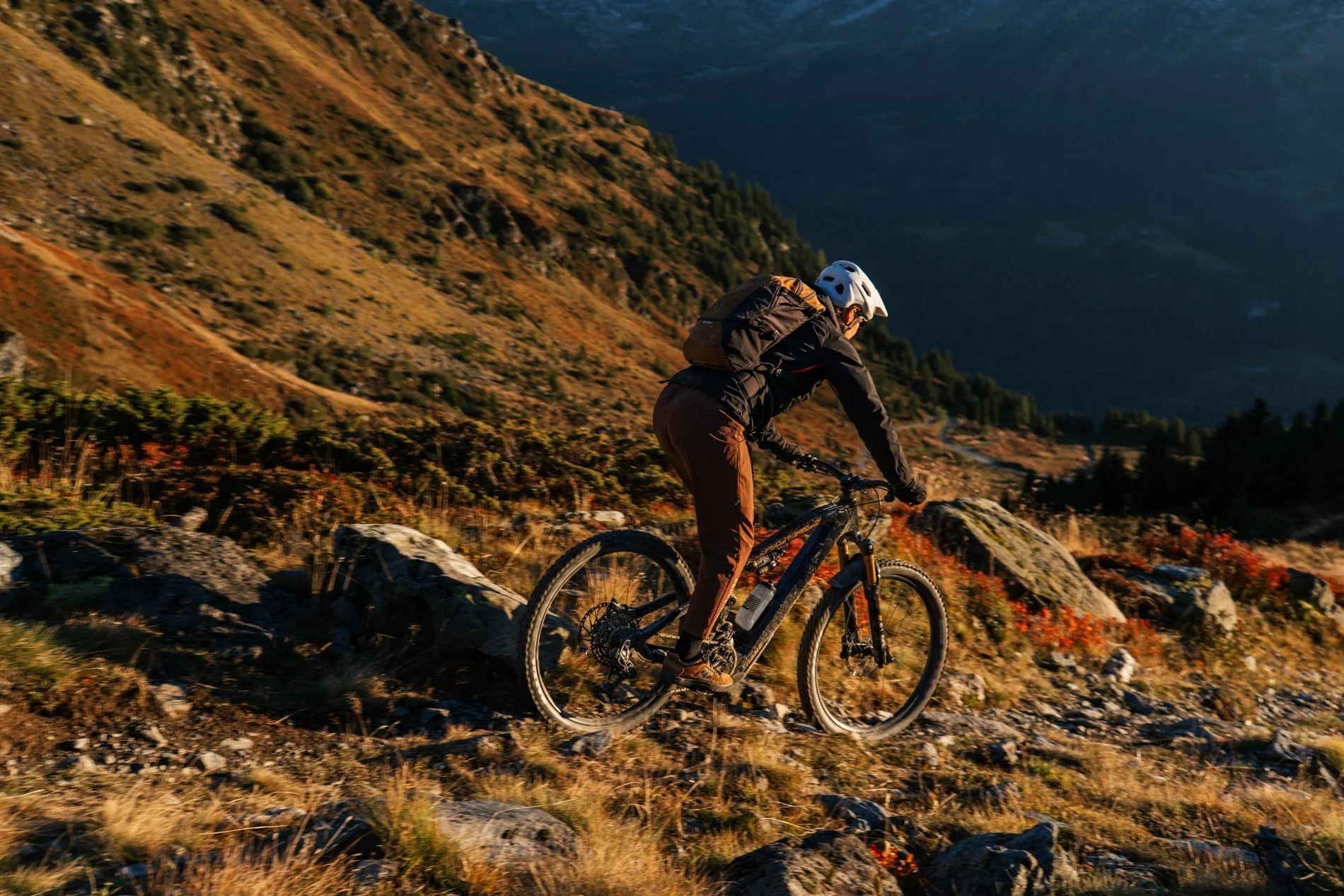 A person riding a mountain bike on rocky terrain through a mountainous landscape during what appears to be late afternoon or early evening, with hills and trees in the background.