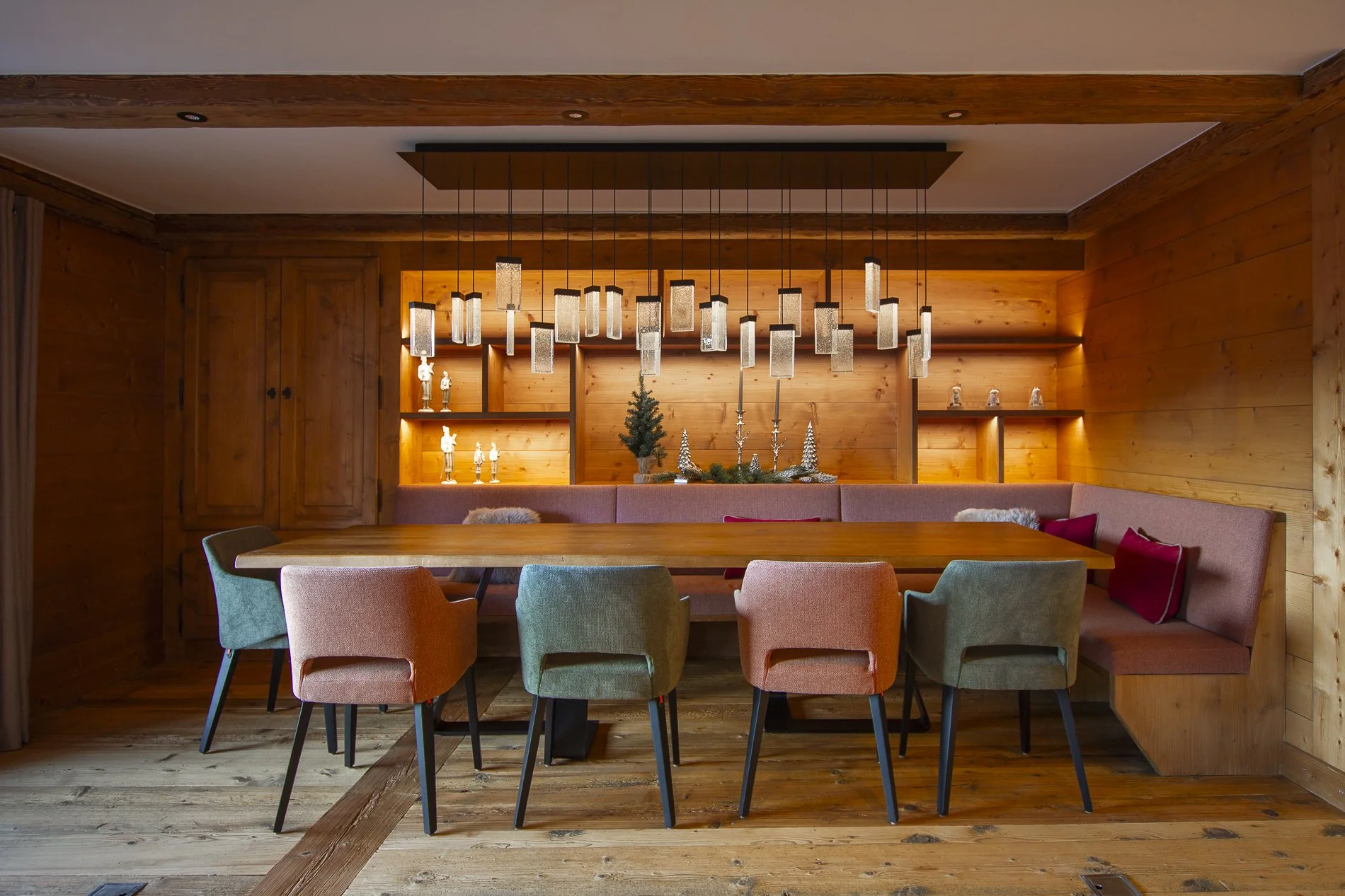 Modern dining room with a wooden table surrounded by pastel-colored chairs and a built-in wooden bench with red and beige cushions. The back wall has wooden shelves decorated with small figurines and candles, with a small Christmas tree and silver or