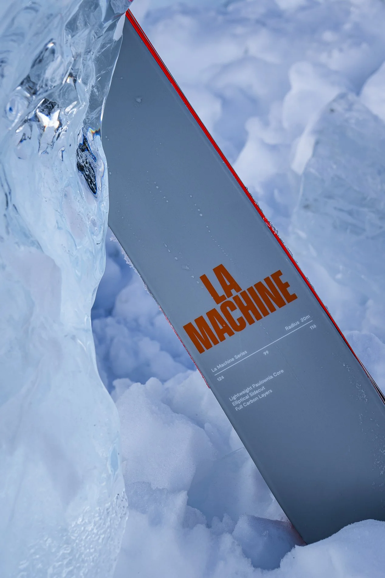 A snowboard named 'La Machine' stuck upright in snow with a block of ice nearby.