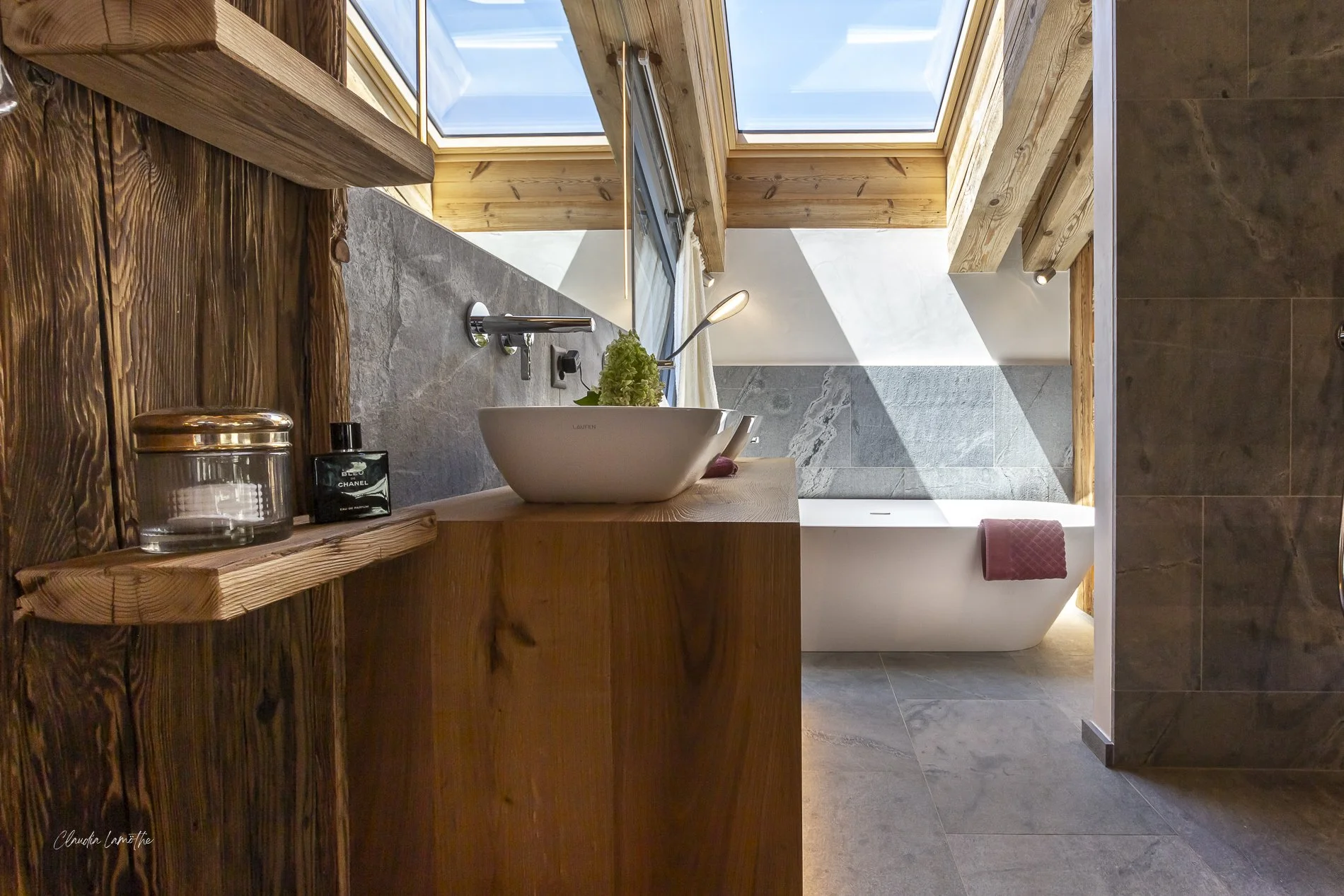 A bathroom with a wooden vanity holding a white vessel sink, a black Chanel perfume bottle, and a glass jar; a ceiling with skylights and a bathtub with a red towel hanging on the side.