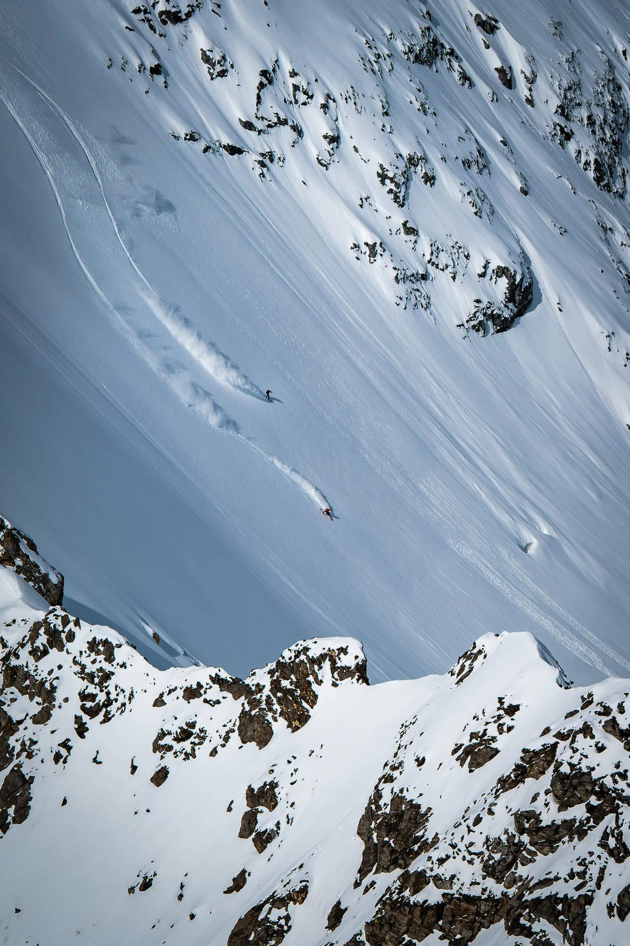 Two skiers descending a snow-covered mountain slope with rocky outcroppings, leaving a trail of snow behind them.