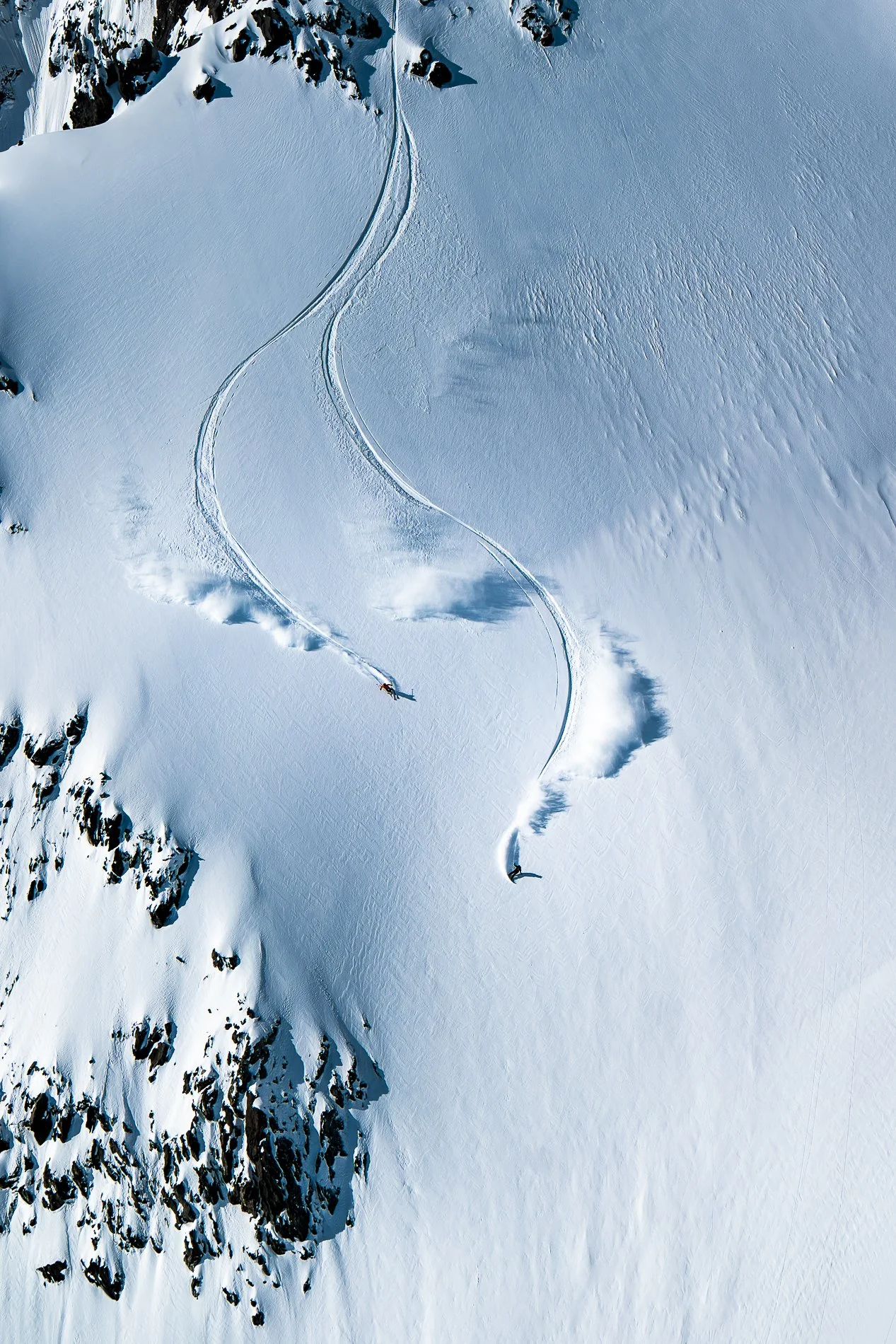 Aerial view of snow-covered mountain slope with ski tracks and climbers near rocky outcrops.
