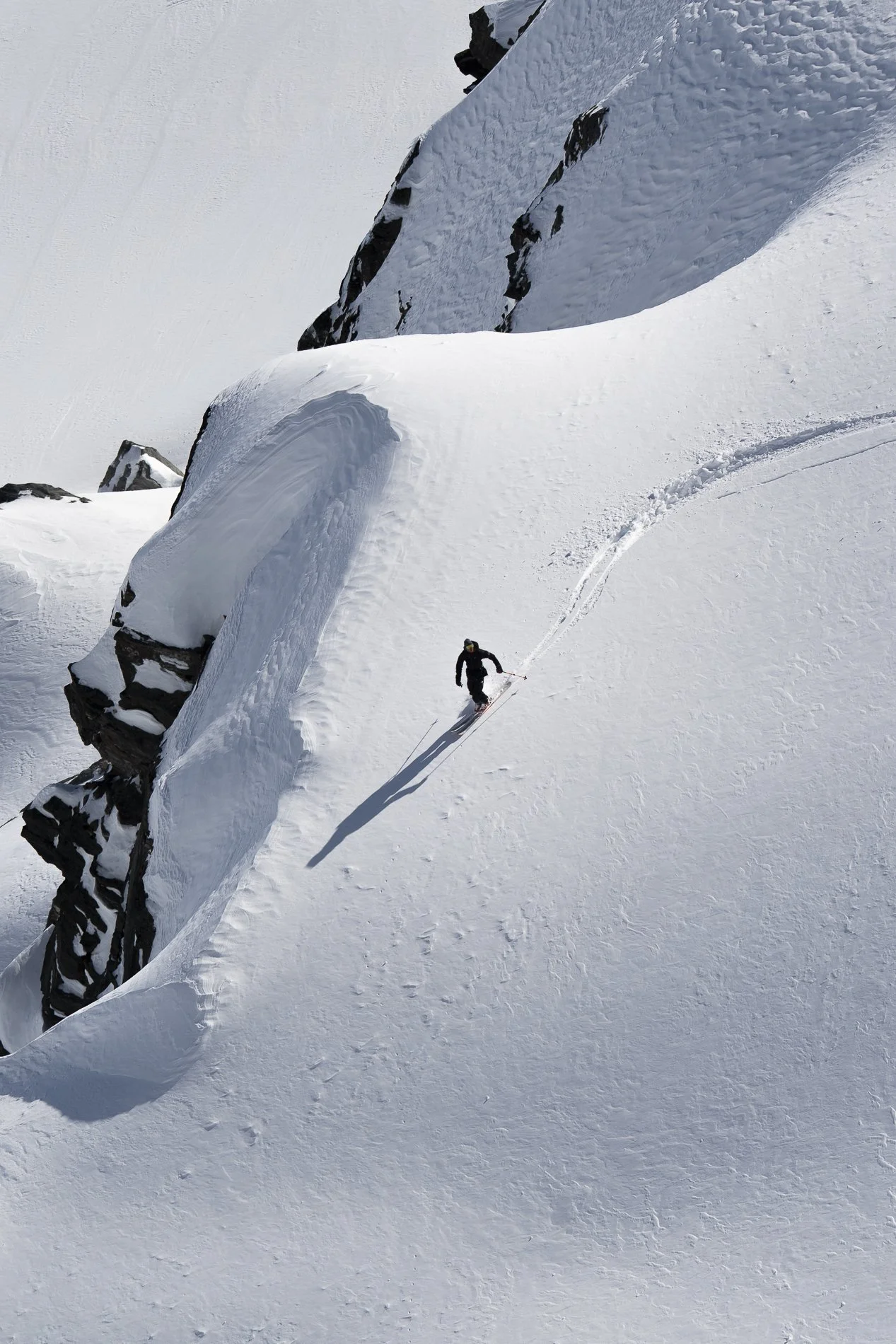 A skier descending a snow-covered slope near large, jagged, snow-covered rocks with a snowy mountain landscape in the background.