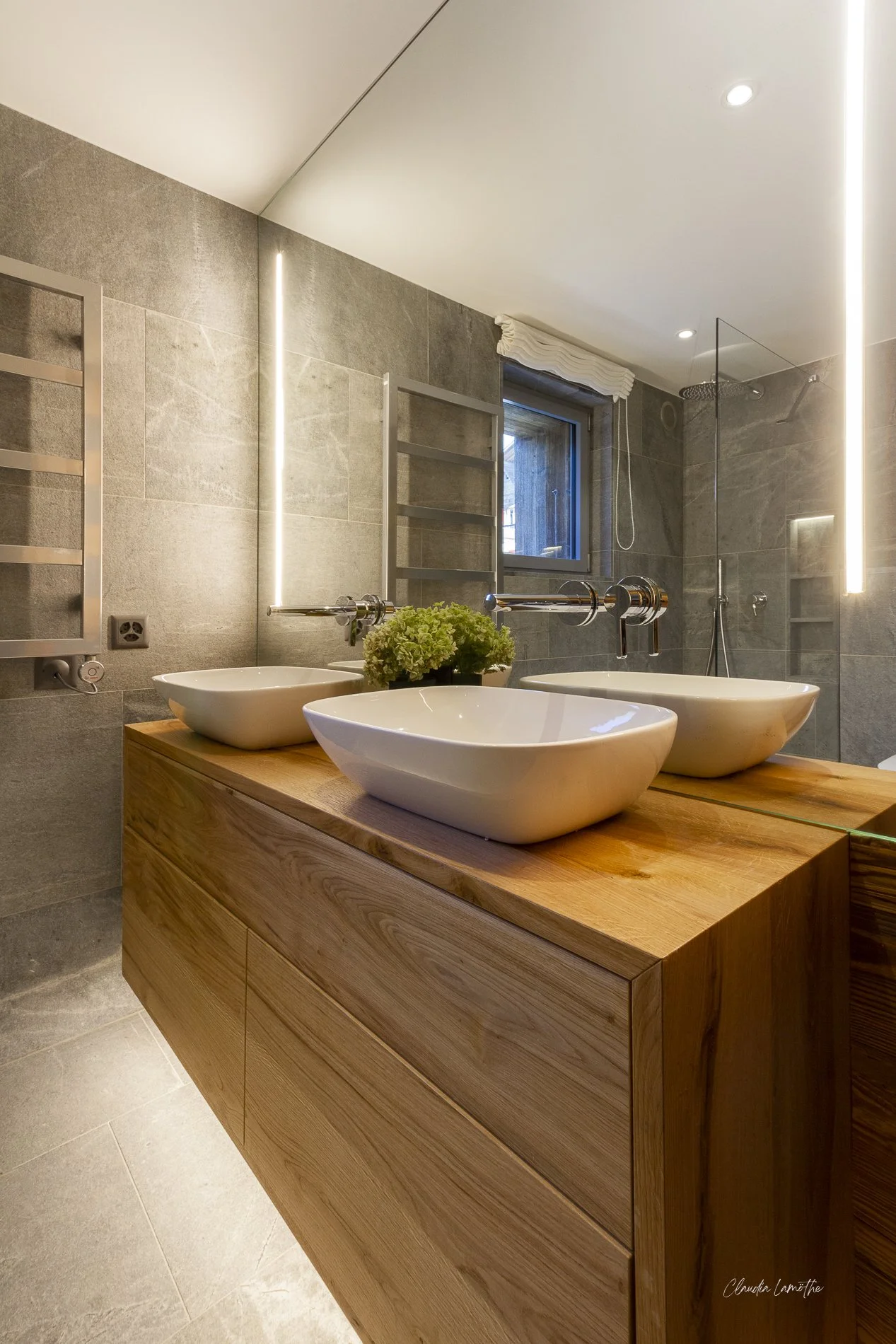 Modern bathroom with a wooden vanity, three vessel sinks, a large mirror, gray tiled walls, a window with a white blind, and a shower with a glass door and rainfall showerhead.