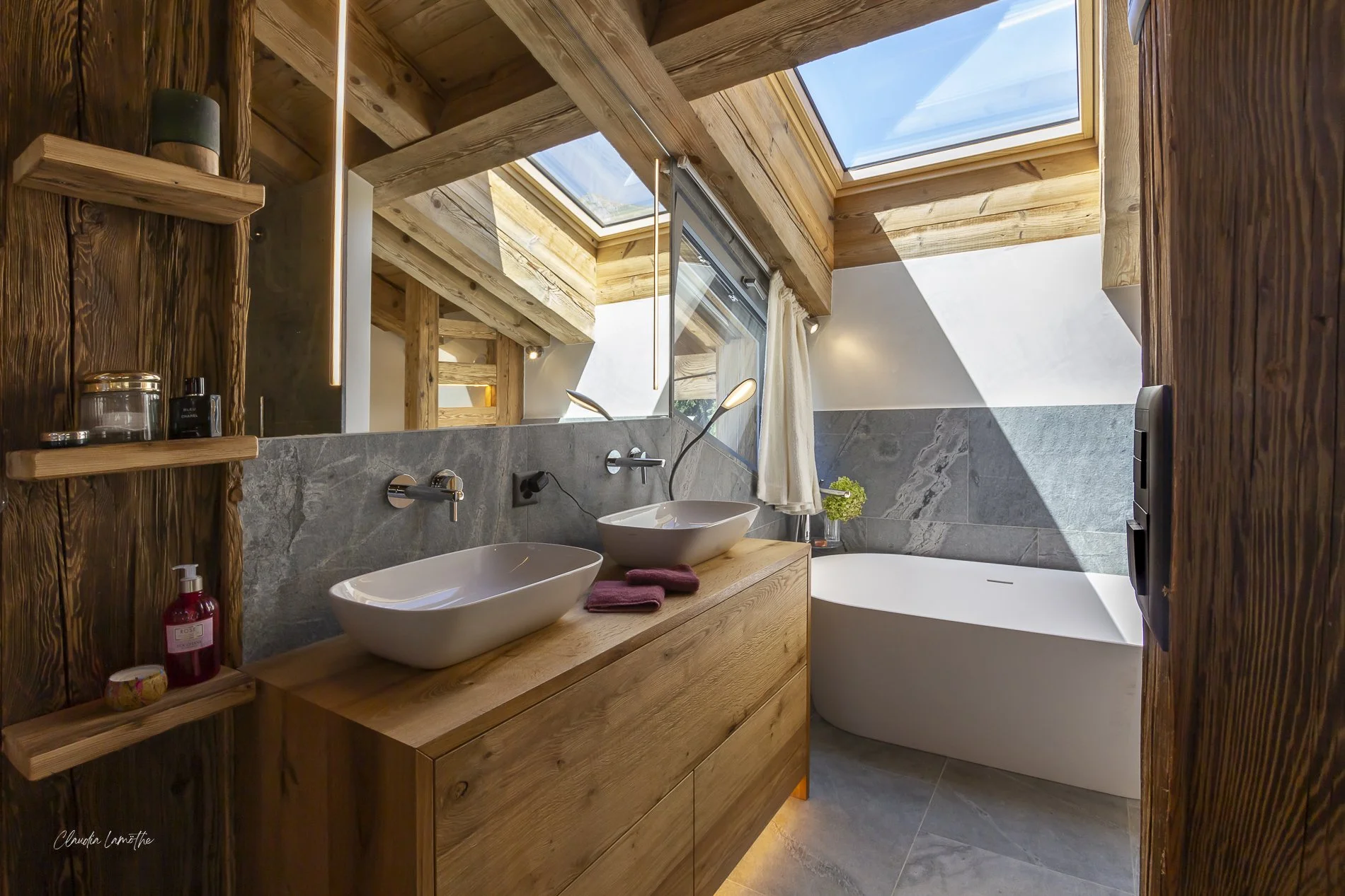 Bathroom with wooden accents, double vessel sinks on a wooden vanity, large skylight windows letting in natural light, and a bathtub under the windows.