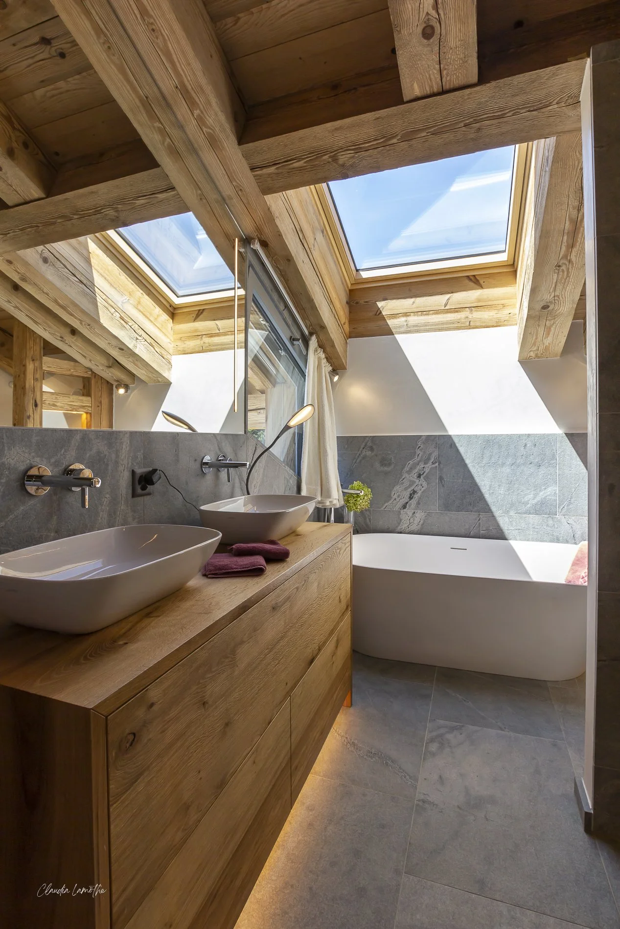 Modern bathroom with two vessel sinks on a wooden vanity, skylight windows, a bathtub, and gray marble floor and wall tiles.