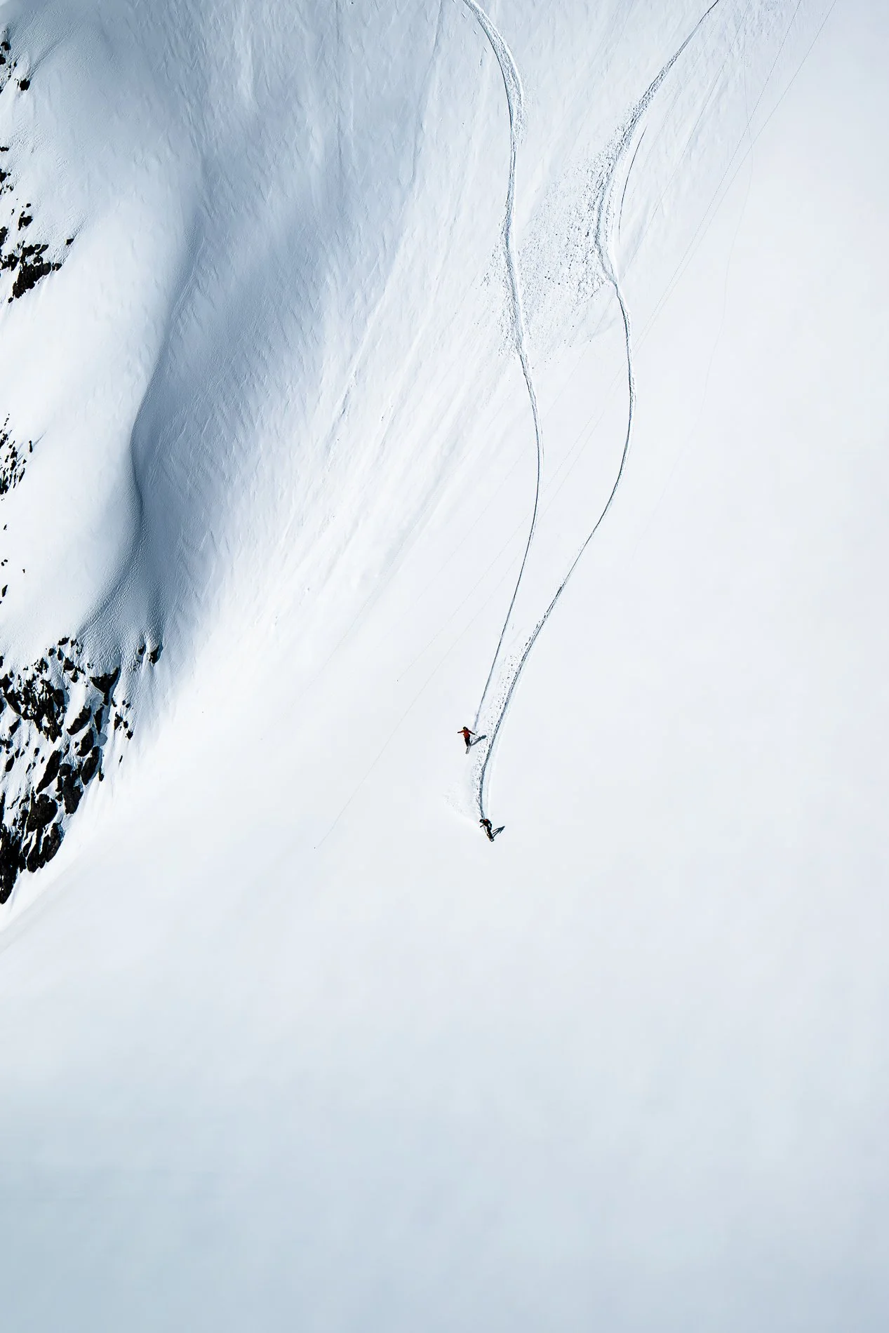 Two skiers moving down a steep, snowy mountain slope with visible ski tracks, surrounded by snow and rock features