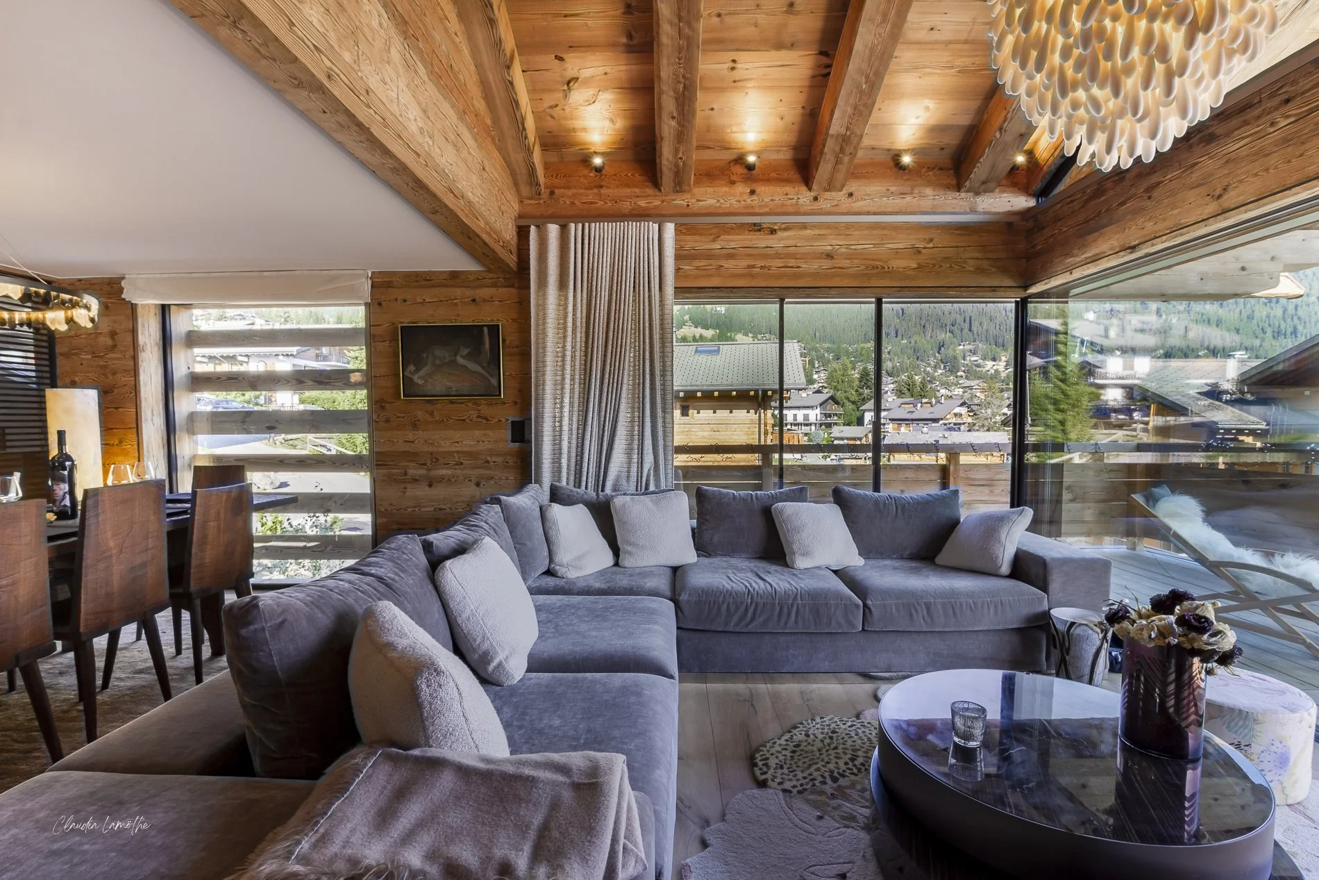 Living room with wooden ceiling beams, large windows showcasing mountain and neighborhood view, plush gray sectional sofa with cushions, round coffee table with floral arrangement and drinks, and a chandelier made of white teardrop-shaped elements.