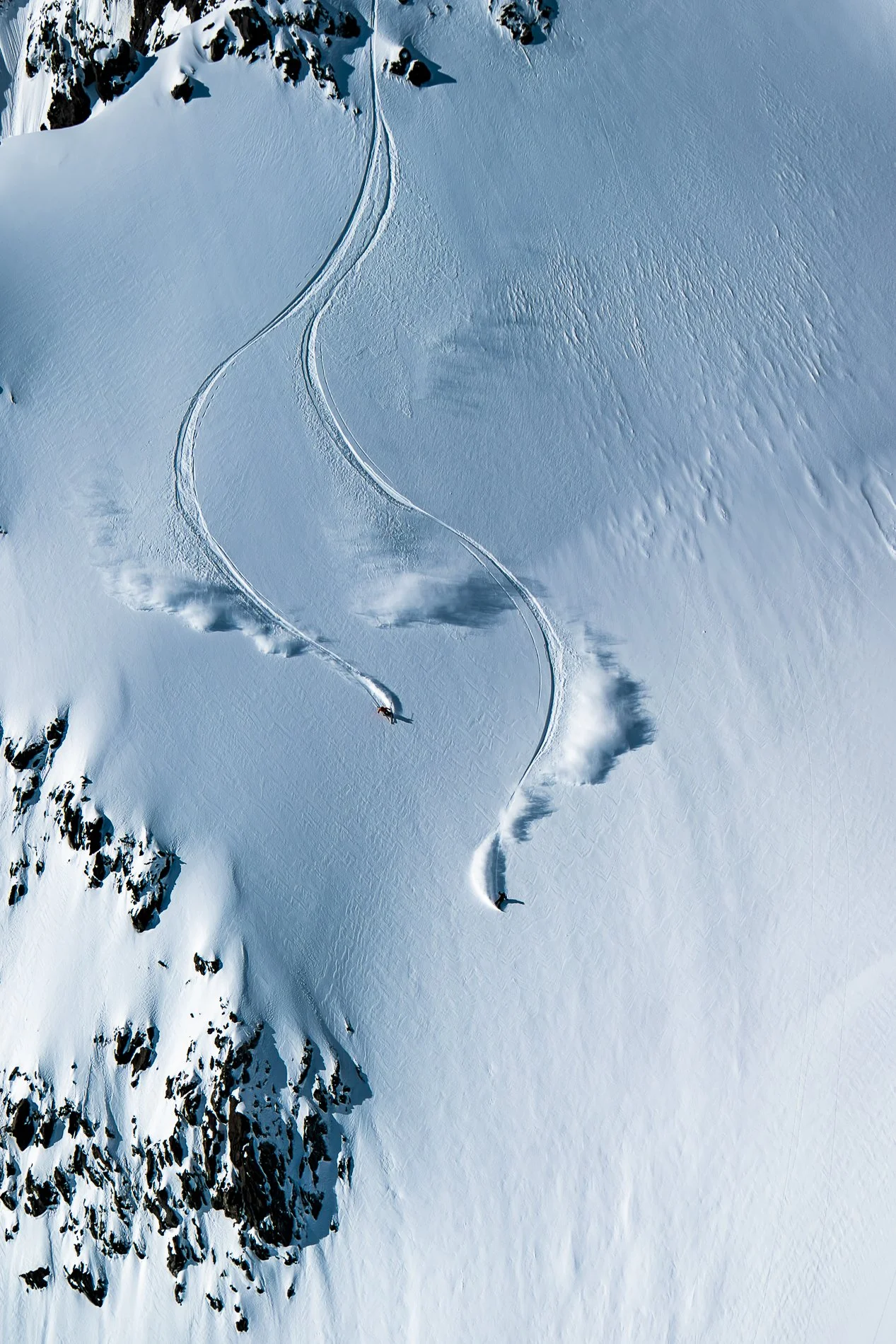Aerial view of snow-covered mountain slopes with two skiers making tracks down the slope.