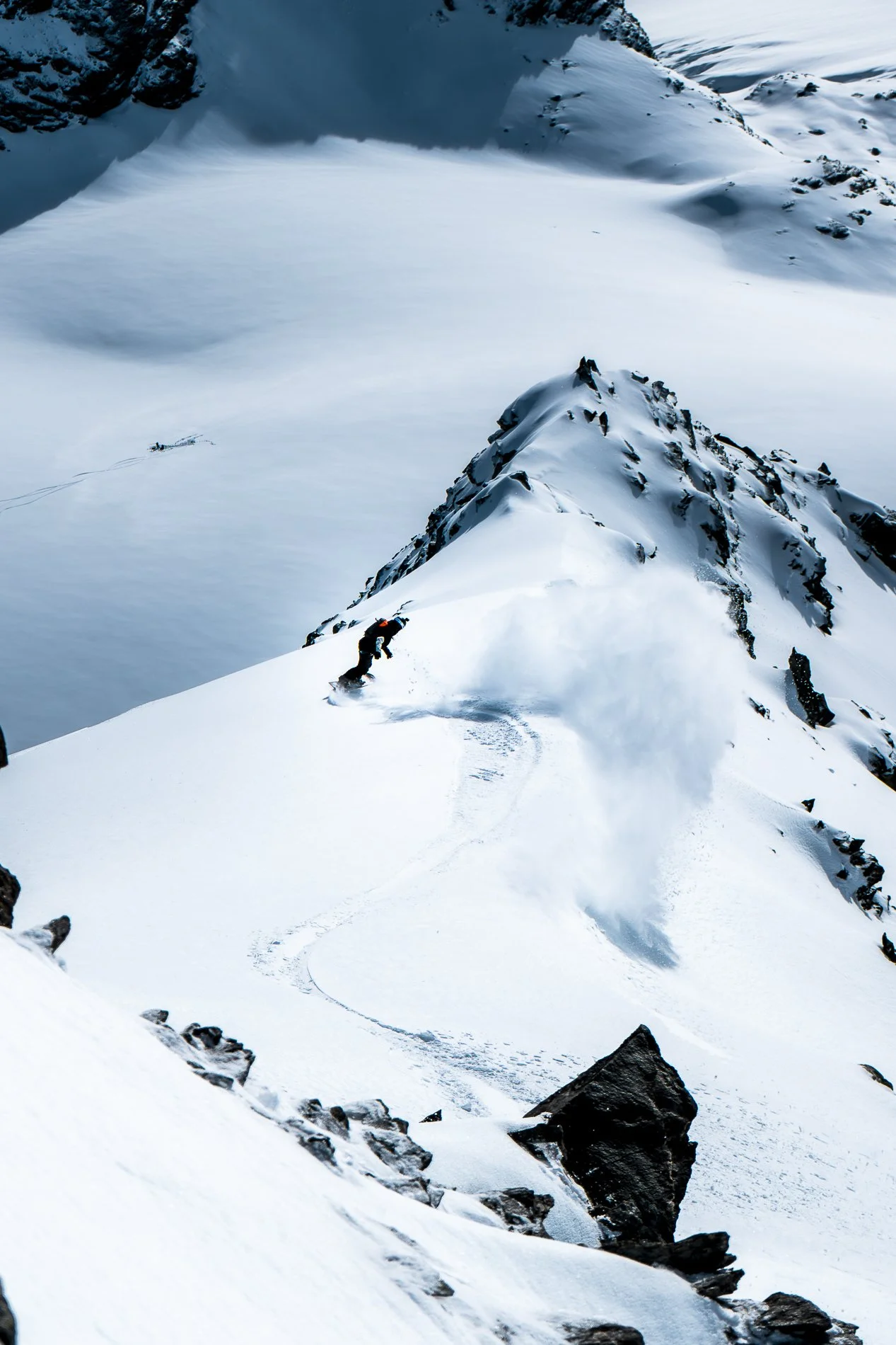 A person snowboarding down a snowy mountain ridge with rocky outcroppings in a remote, mountainous landscape.