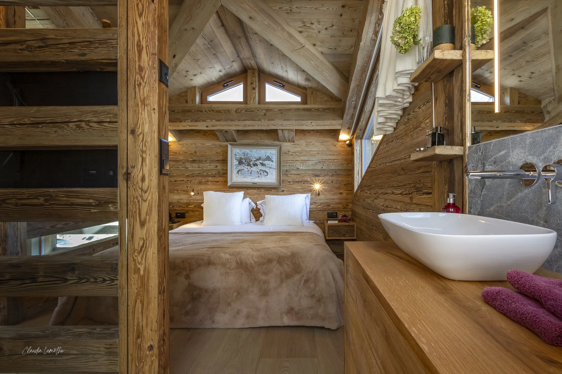 A cozy wooden cabin bedroom with a vaulted ceiling, a bed with white pillows, a framed winter scene painting above, and a small sink with bathroom essentials on a wooden counter.