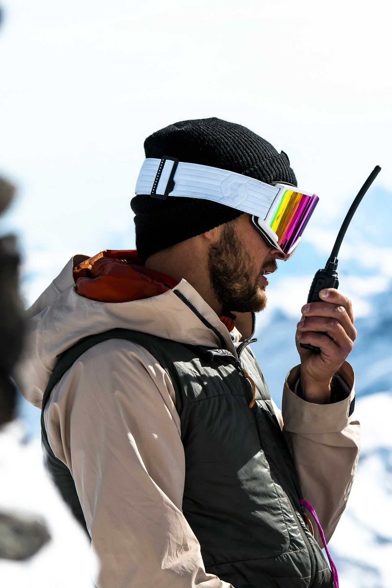 A man in winter gear, including a black knit hat, beige jacket, and black vest, holds a walkie-talkie while outdoors with snow and mountains in the background.