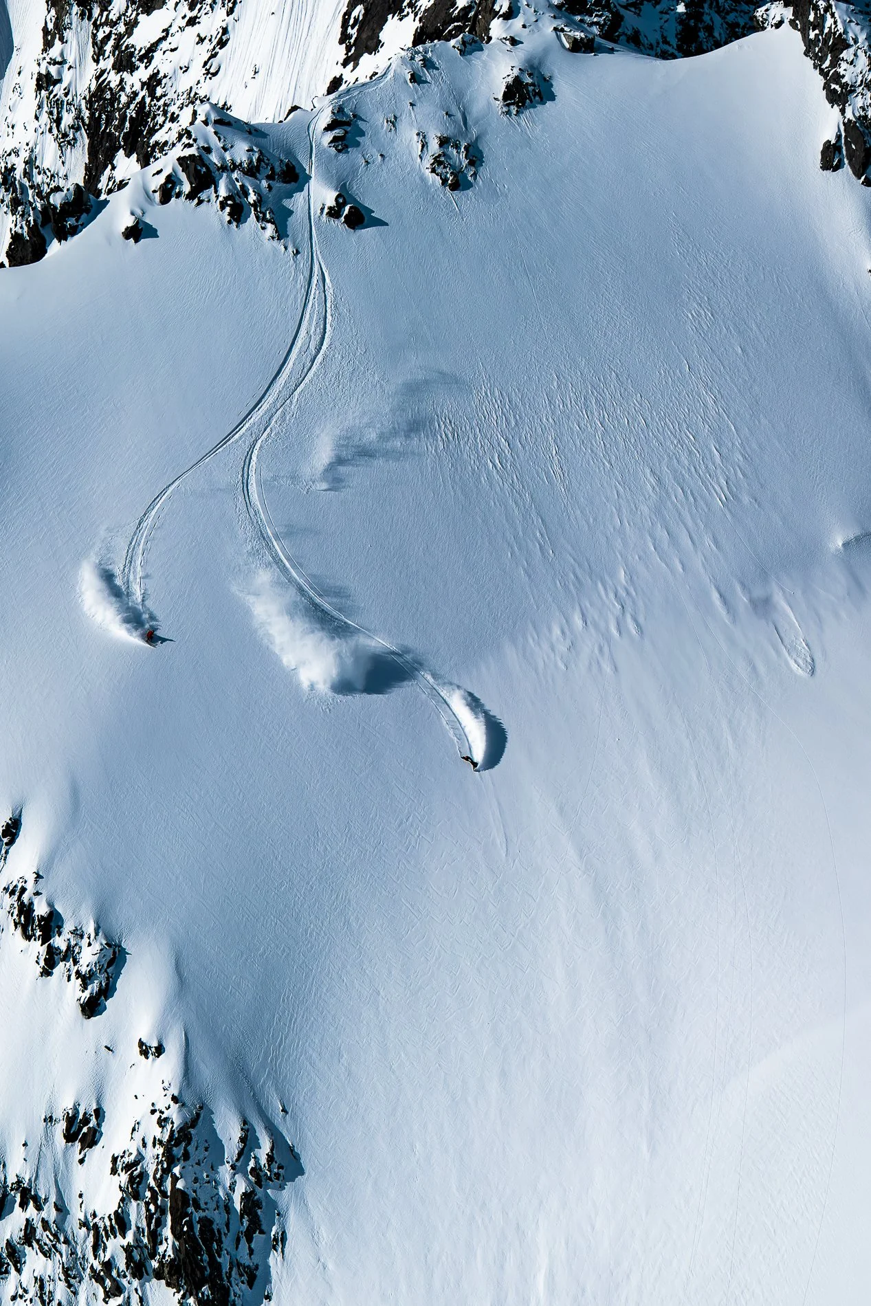 Aerial view of a snow-covered mountain slope with two skiers making turns, creating ski tracks on the snow. The slope is adjacent to rocky mountain outcrops.