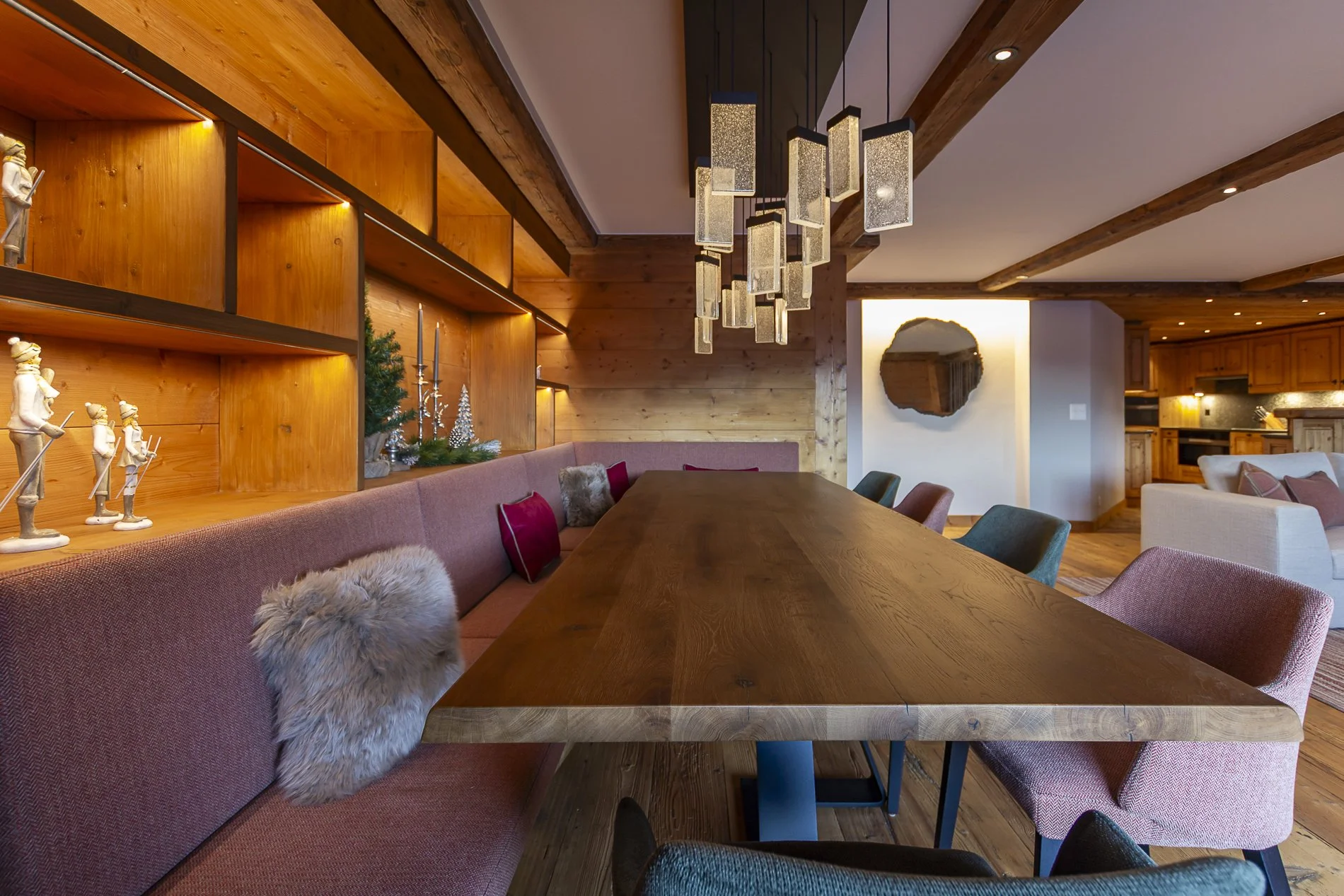 Modern dining room with wooden walls and ceiling, a long wooden table surrounded by pink and teal chairs, a large wooden shelf with decorative figurines, Christmas decorations, and a contemporary chandelier.