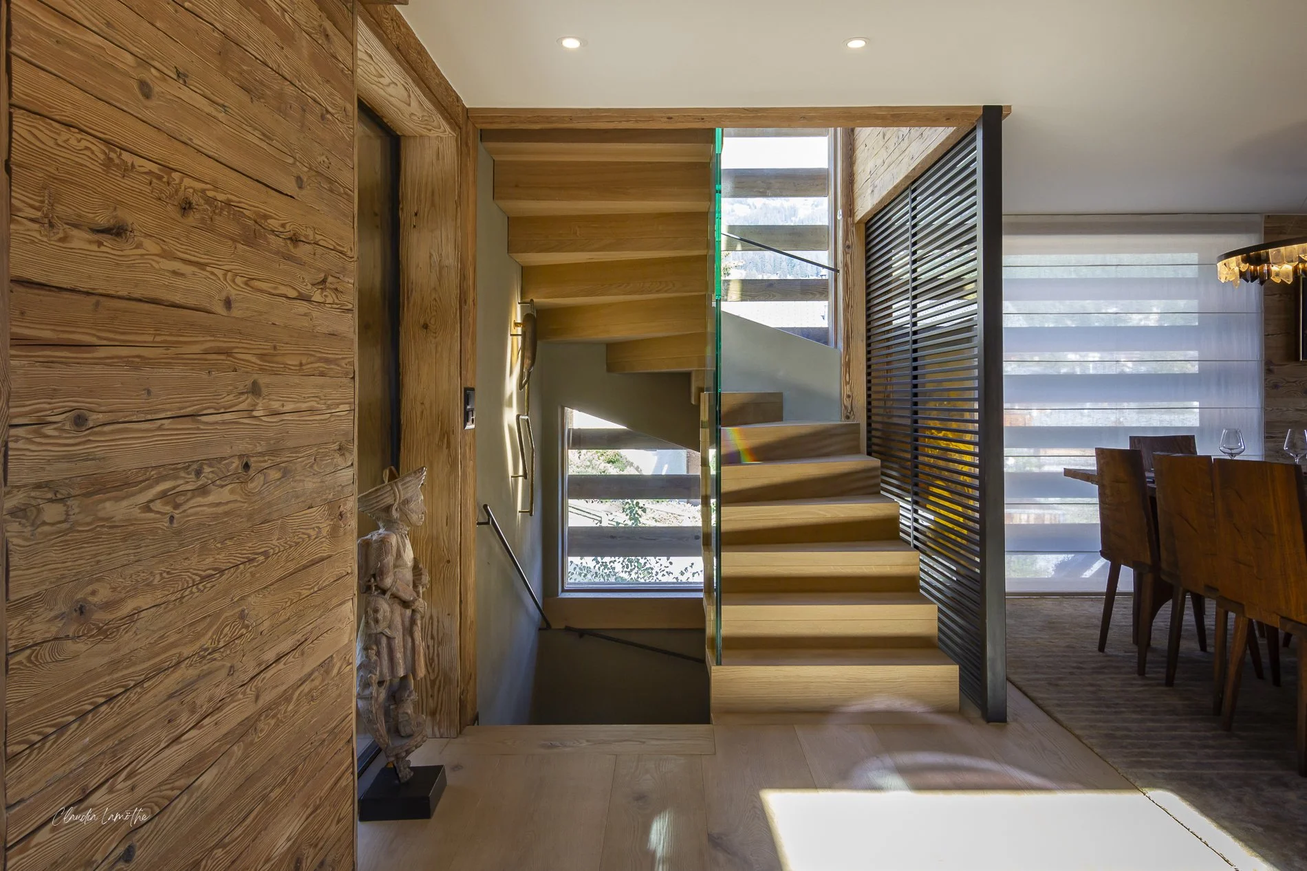Interior view of a modern wooden staircase with glass railing, wood paneling, and large windows allowing natural light into the space.