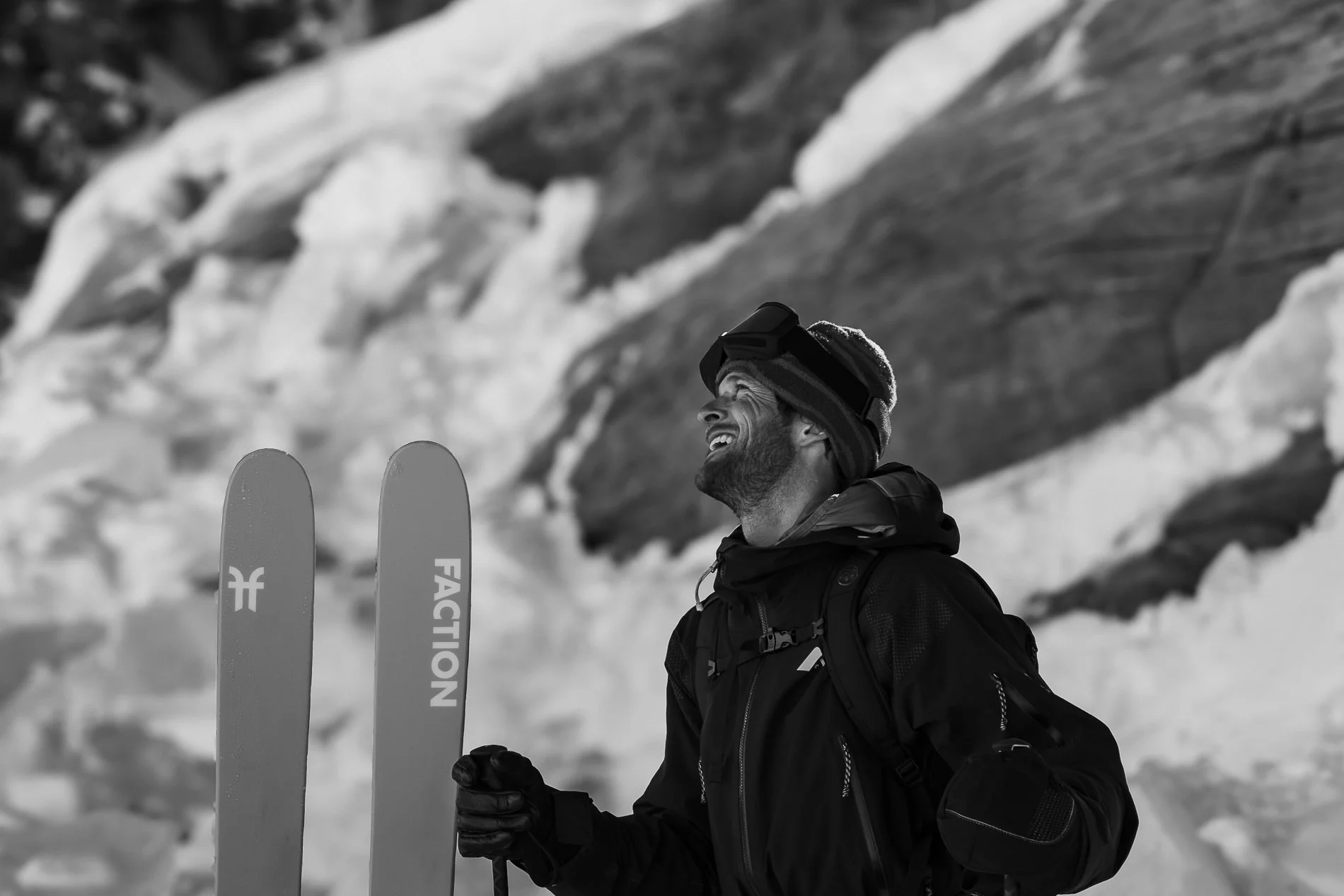 A man smiling in snow with skis in hand, wearing a black jacket and goggles on his head, against a snowy mountain background.