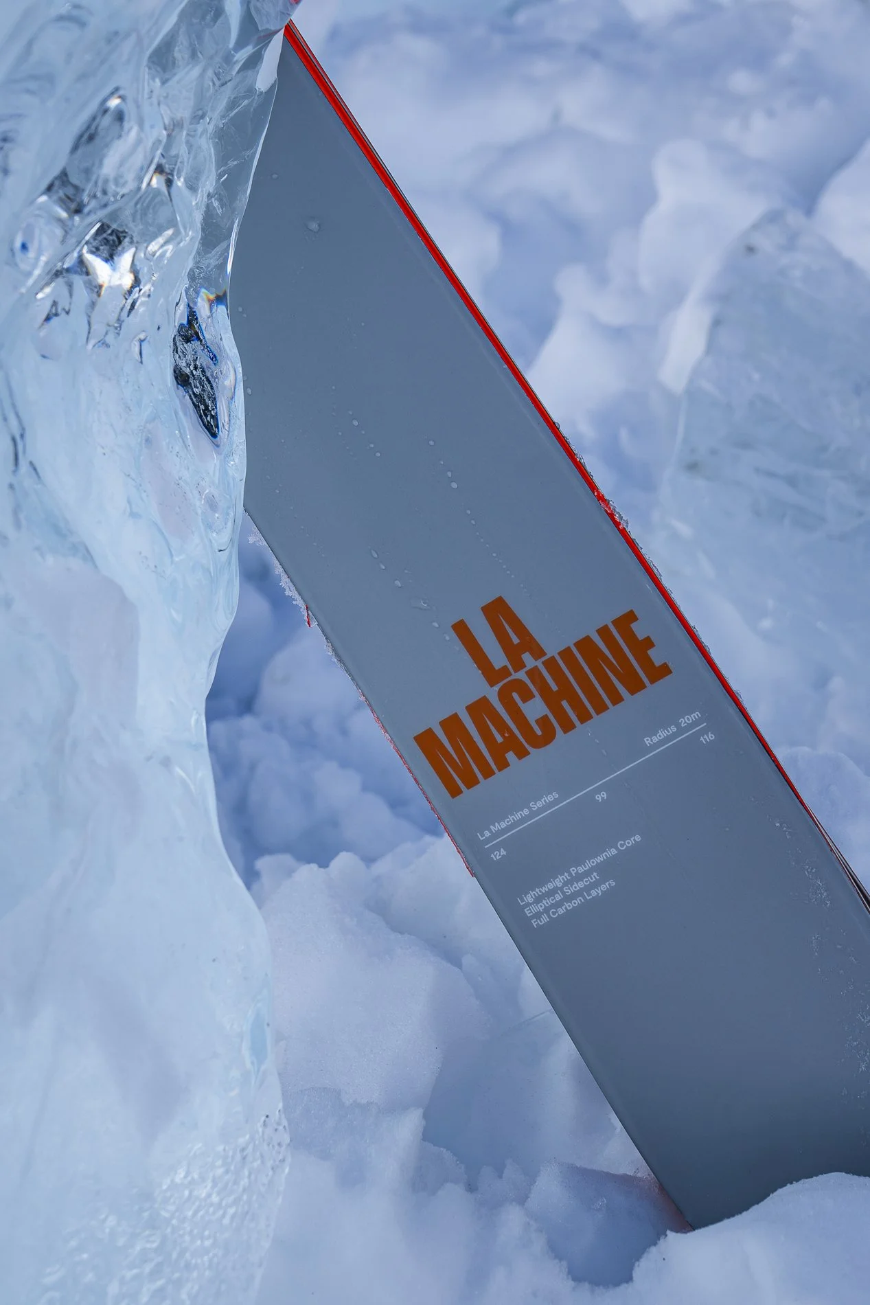 Close-up of a skateboard called 'La Machine' partially embedded in snow, with reeds or ice formations in the background.