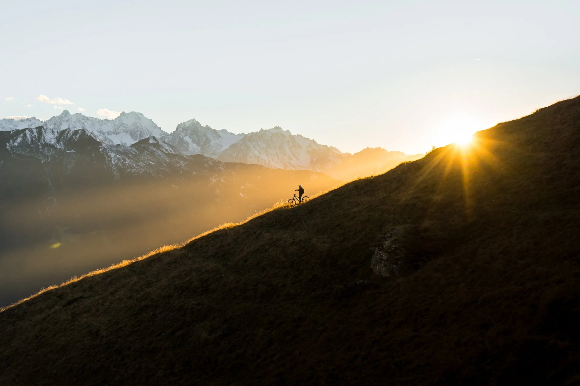 A person with a bicycle standing on a grassy hillside at sunrise or sunset, with snow-capped mountains in the background and sunlight casting rays over the hillside.