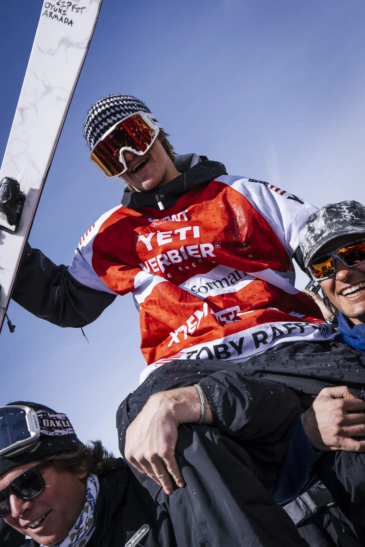 Group of skiers celebrating after a run, wearing ski gear and sunglasses, with a clear blue sky in the background.