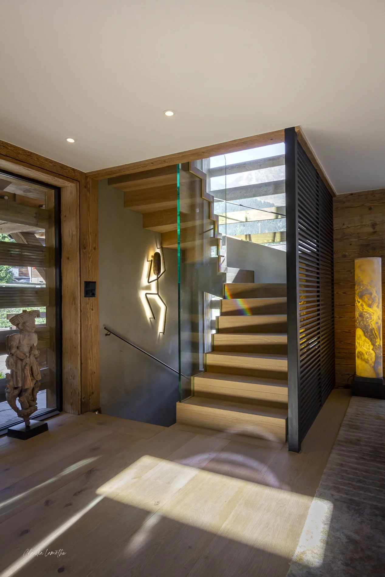 Interior view of a modern wooden staircase with glass railing, overlooking a room with wooden floors and walls, decorated with a lit yellow lamp and a sculpture, in a house with large windows showing greenery outside.