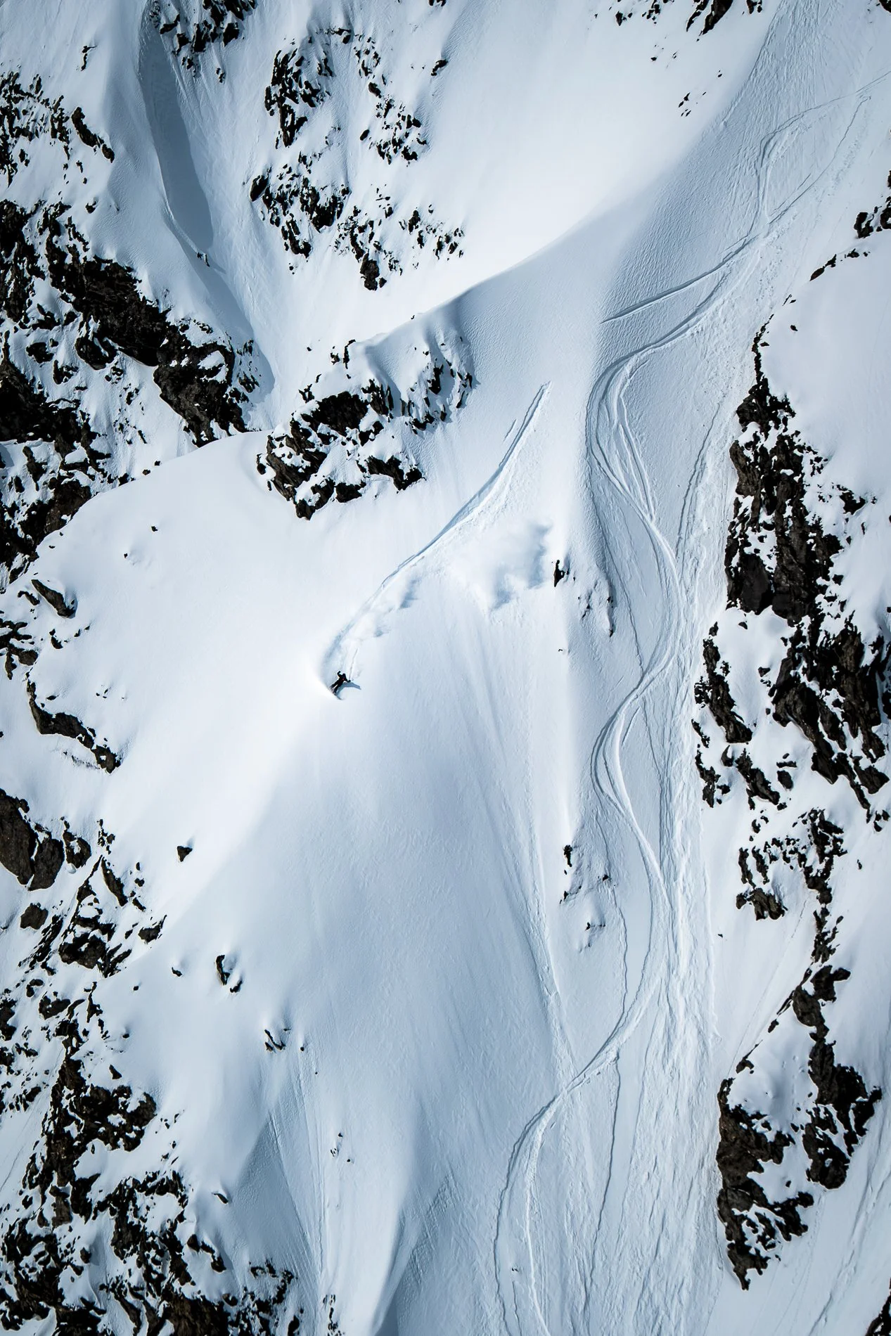 Aerial view of a snow-covered mountain landscape with ski tracks and rocky outcroppings.
