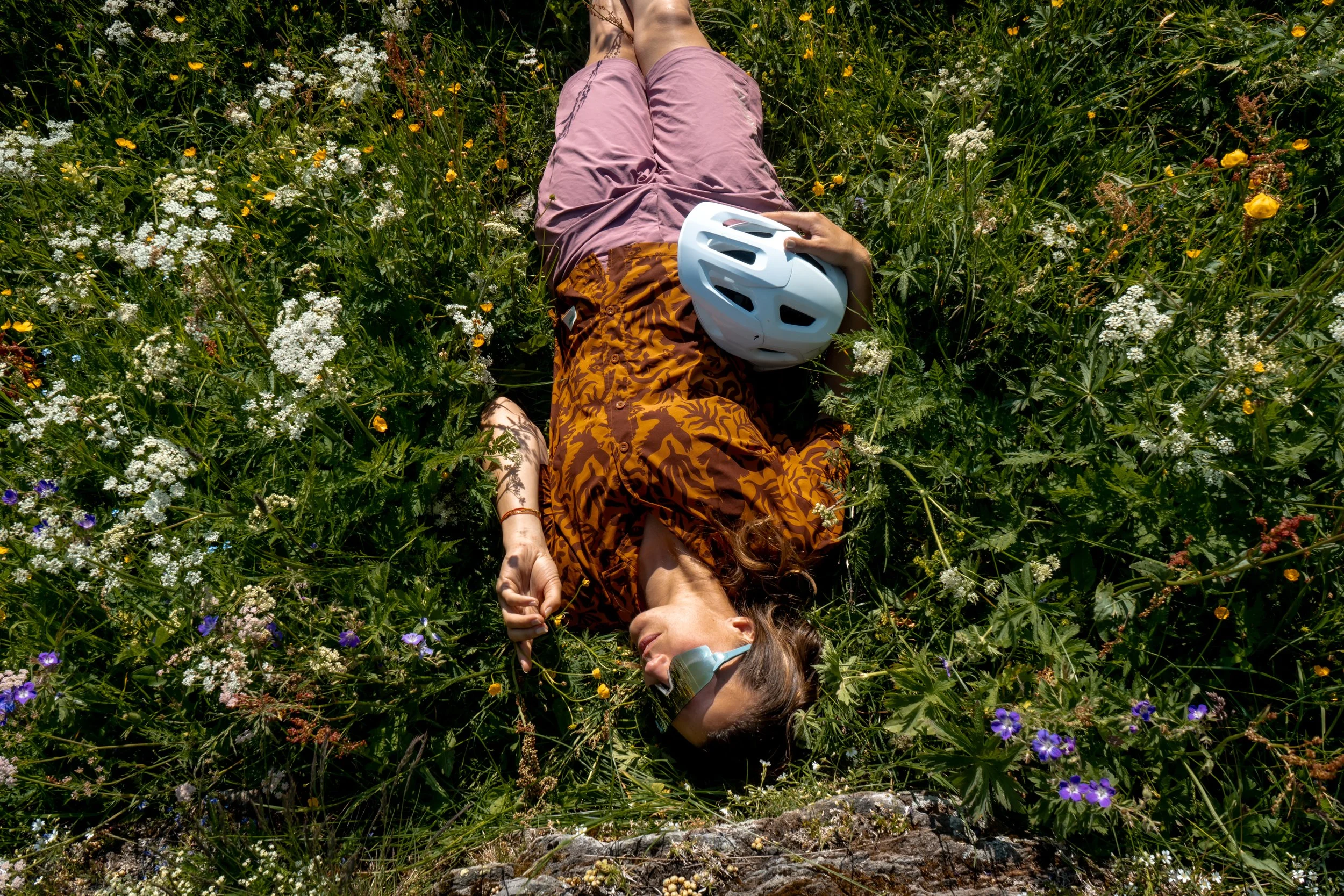 A person lying on their back in a field of wildflowers, wearing a patterned orange and brown shirt, pink shorts, sunglasses, and a white helmet. Their eyes are closed, and they are surrounded by green plants and colorful flowers.