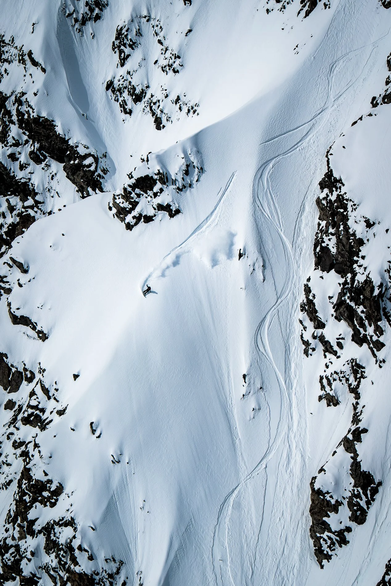 Aerial view of a snow-covered mountain slope with ski tracks and rocky outcroppings.