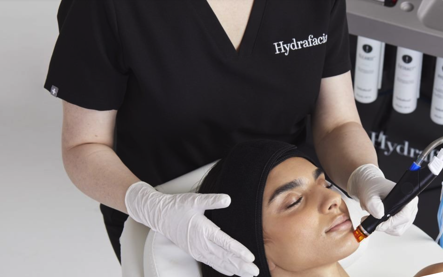 A woman lying down with a headband, receiving a skincare treatment from a professional wearing gloves.